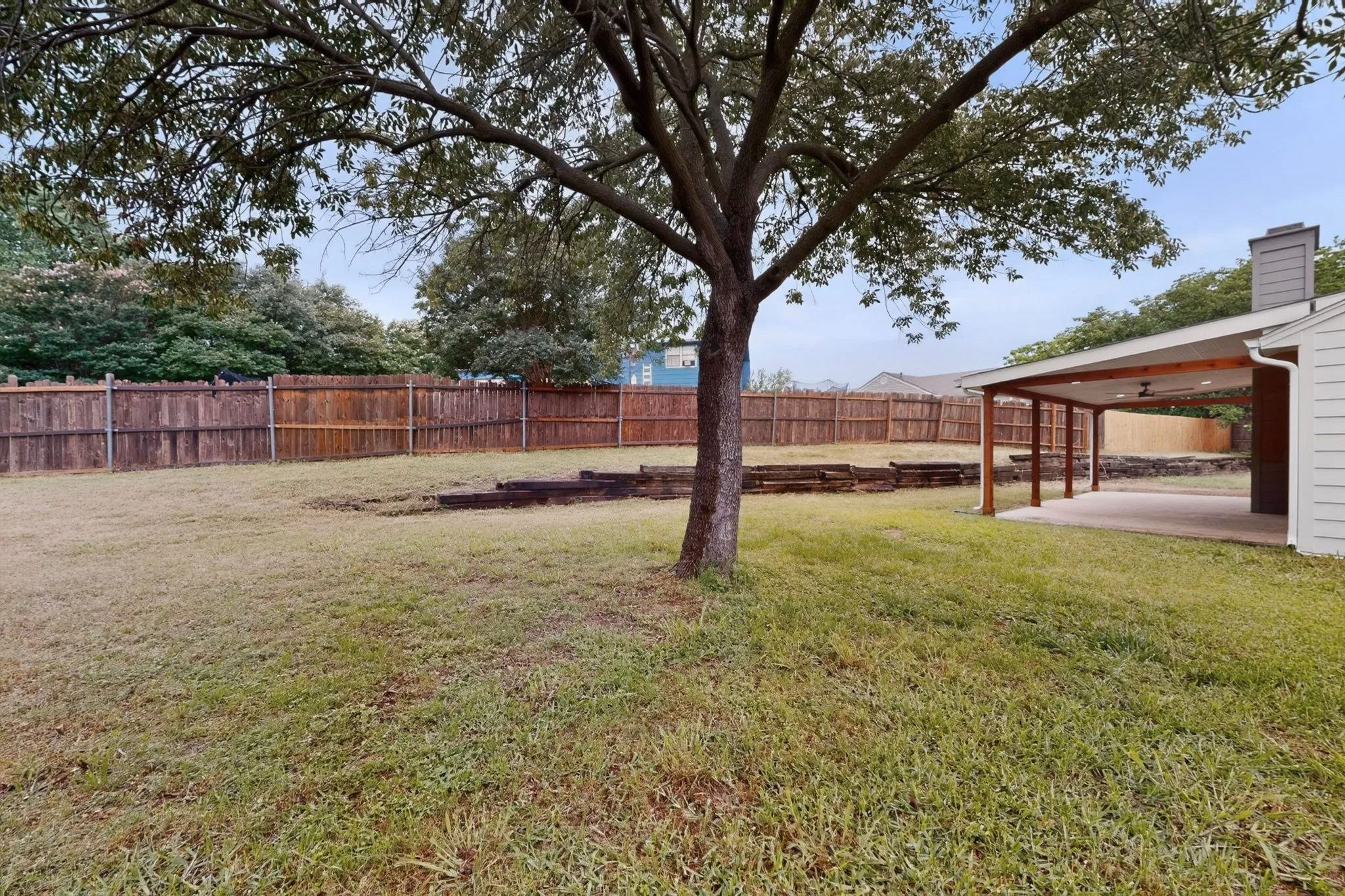Expansive backyard featuring a mature tree, wood privacy fencing, and a covered patio with exposed beams and recessed lighting