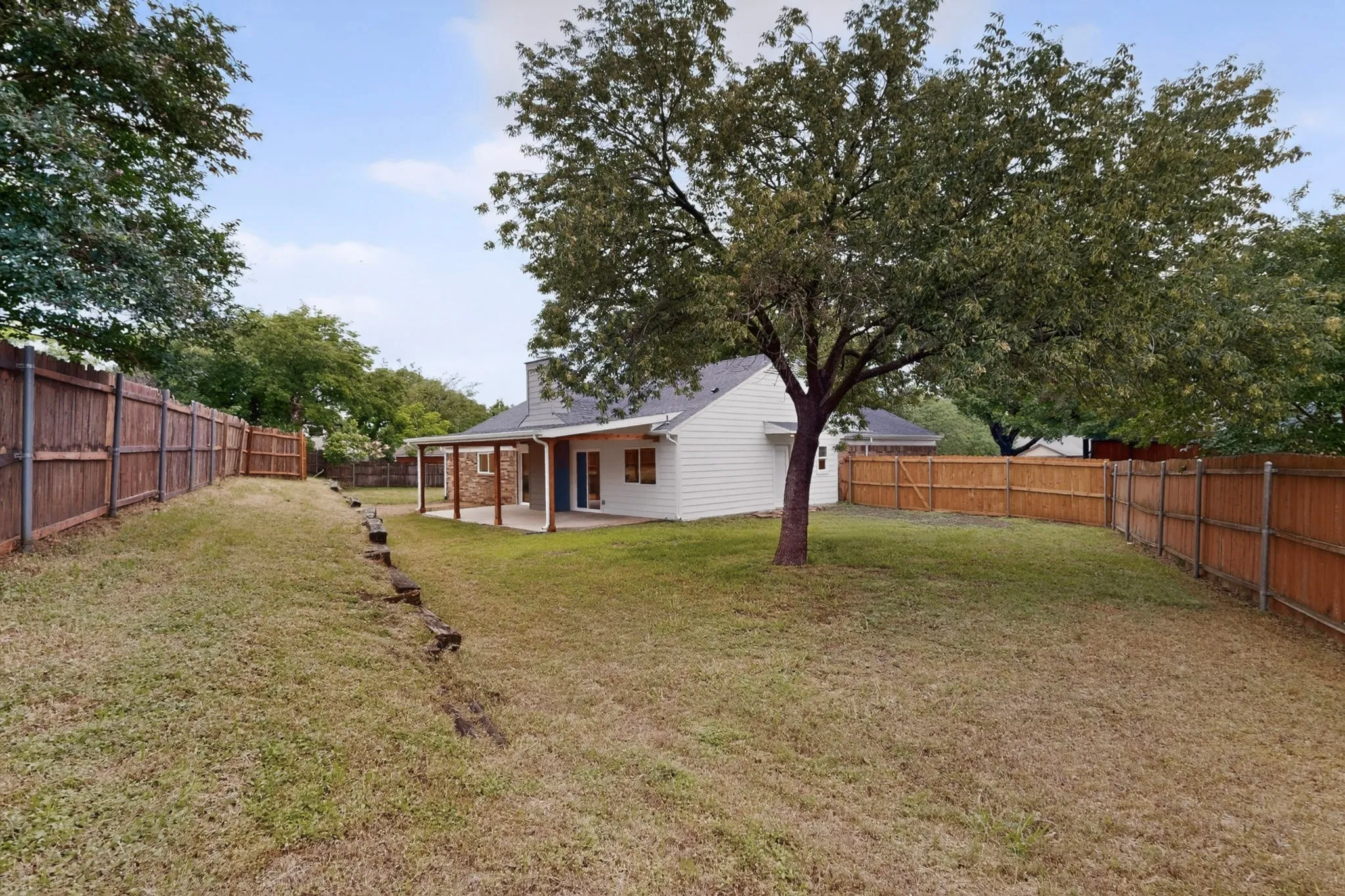 Spacious backyard featuring an established shade tree, a covered patio with exposed beams, a stone accent wall, and perimeter wood fencing