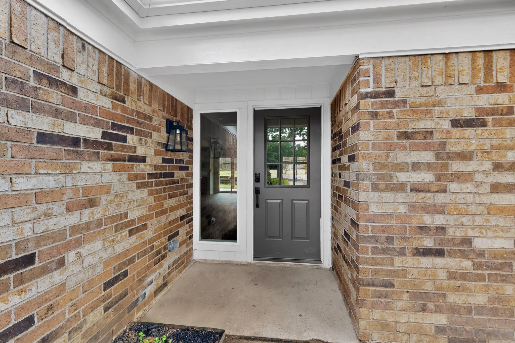 Entryway featuring a multi-toned brick facade, a dark gray door with glass lites, a matching fixed sidelite, and an exterior wall lantern