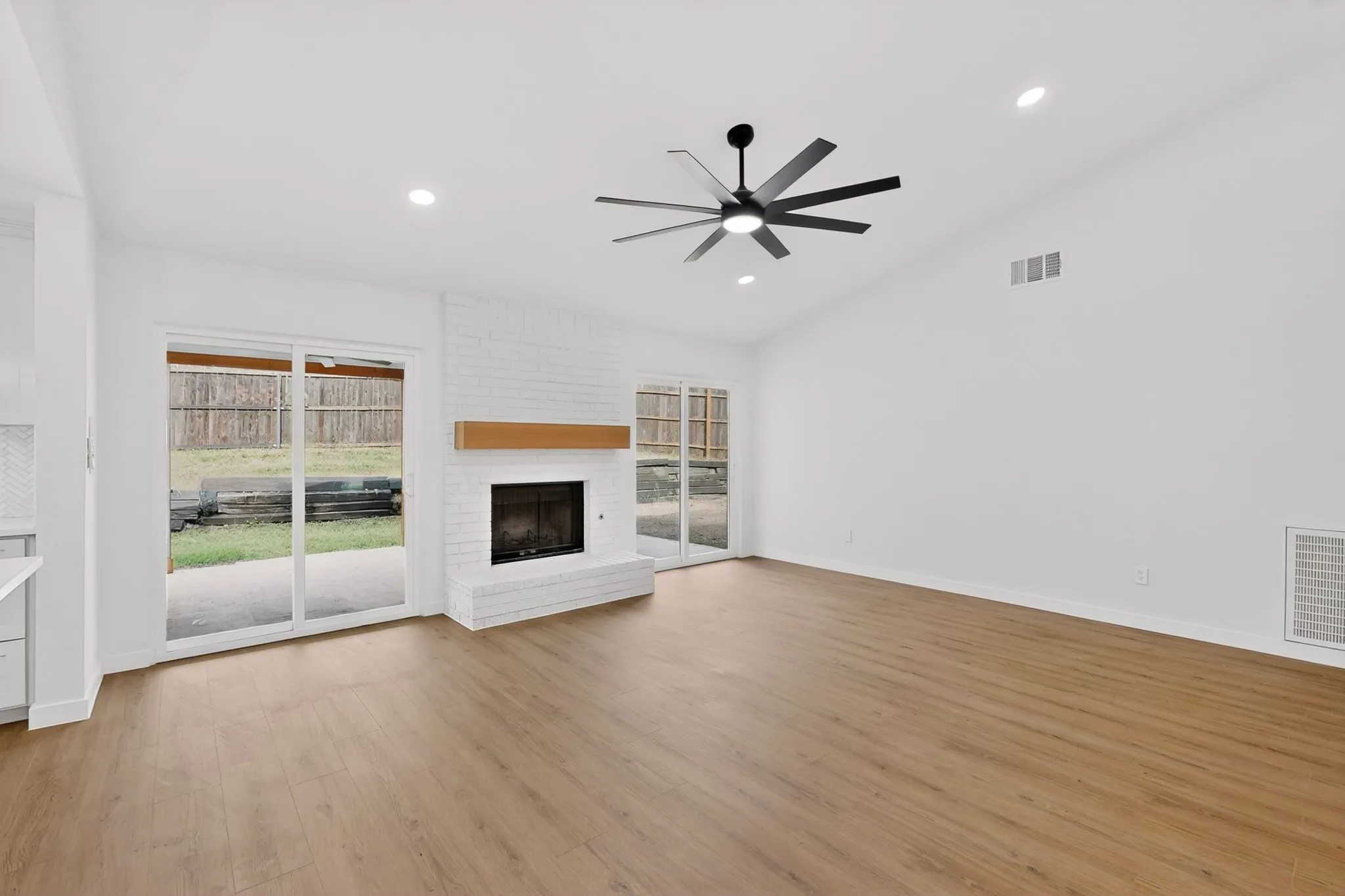 Spacious living area featuring wood-finish flooring, a white-painted brick fireplace with wood mantel, vaulted ceiling with recessed lighting, and a modern ceiling fan