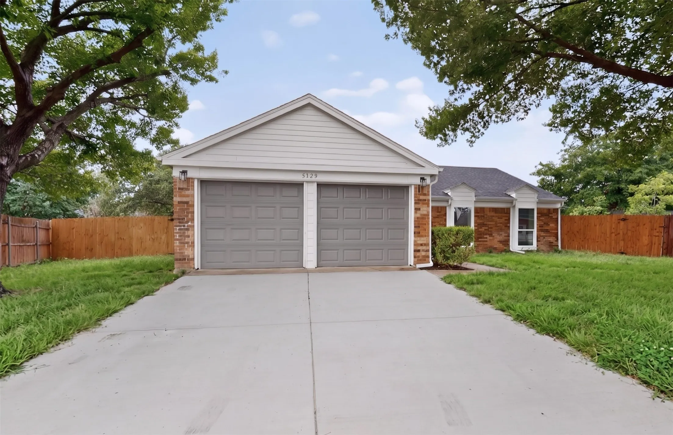 Brick exterior featuring a two-car garage with panel doors, a concrete driveway, a gabled roofline, and established trees