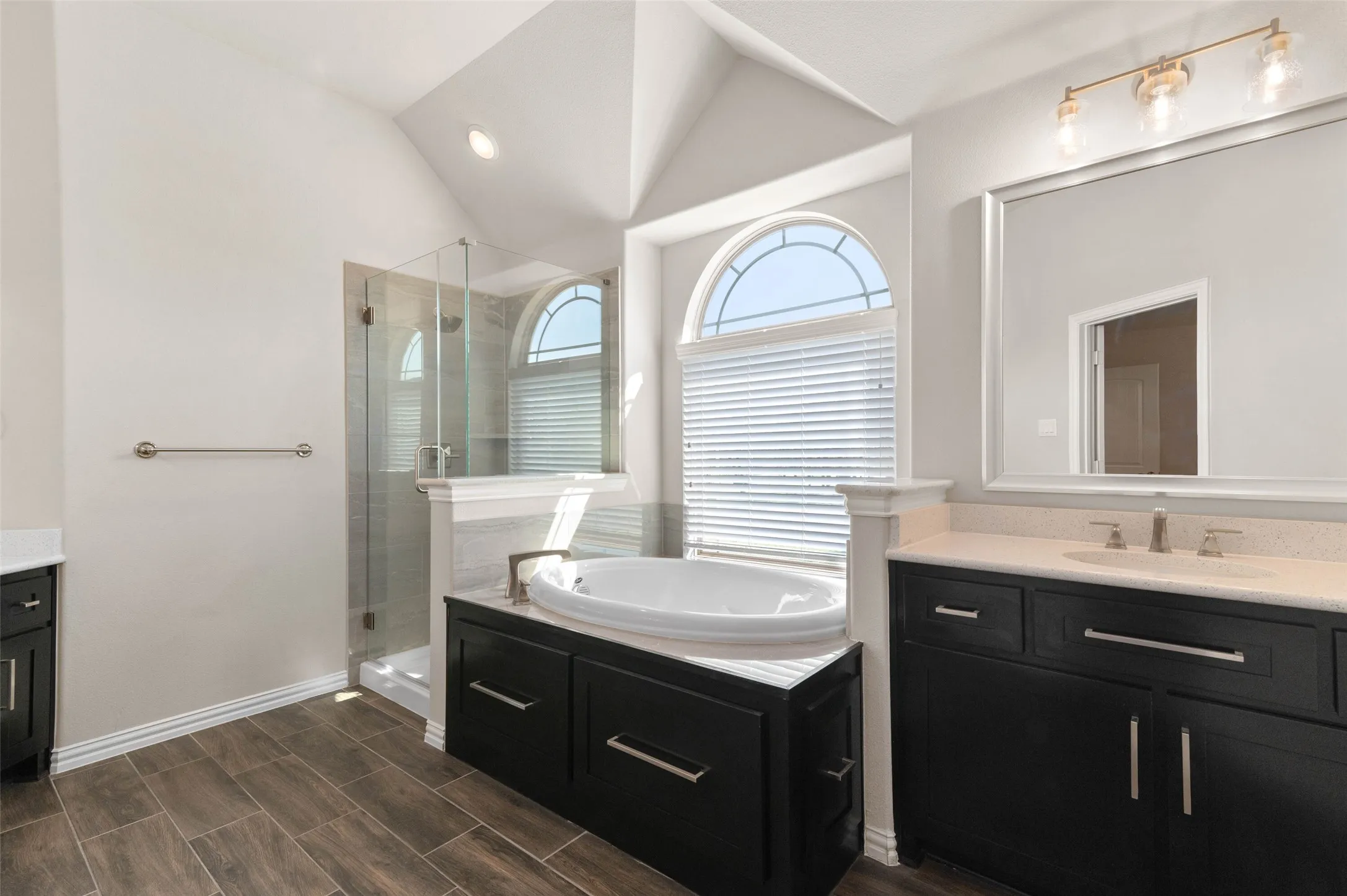 Secondary primary Bathroom featuring a built-in soaking tub, a glass-enclosed shower, dual dark wood vanities with light countertops, and wood-finish flooring