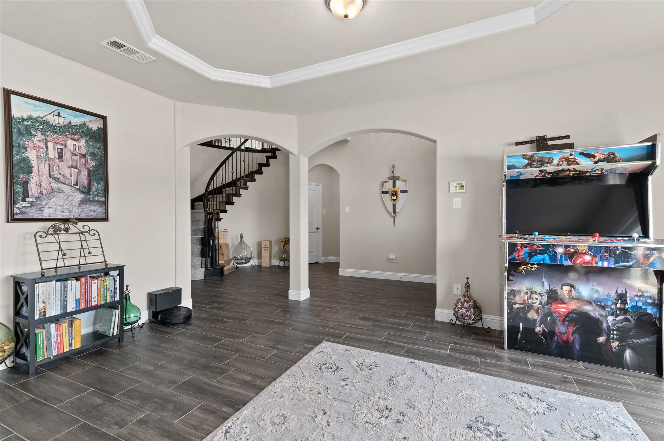 Expansive open-concept living space featuring wood-finish flooring, arched doorways, a tray ceiling with crown molding, and a prominent curved staircase with dark wood handrails and wrought iron balusters