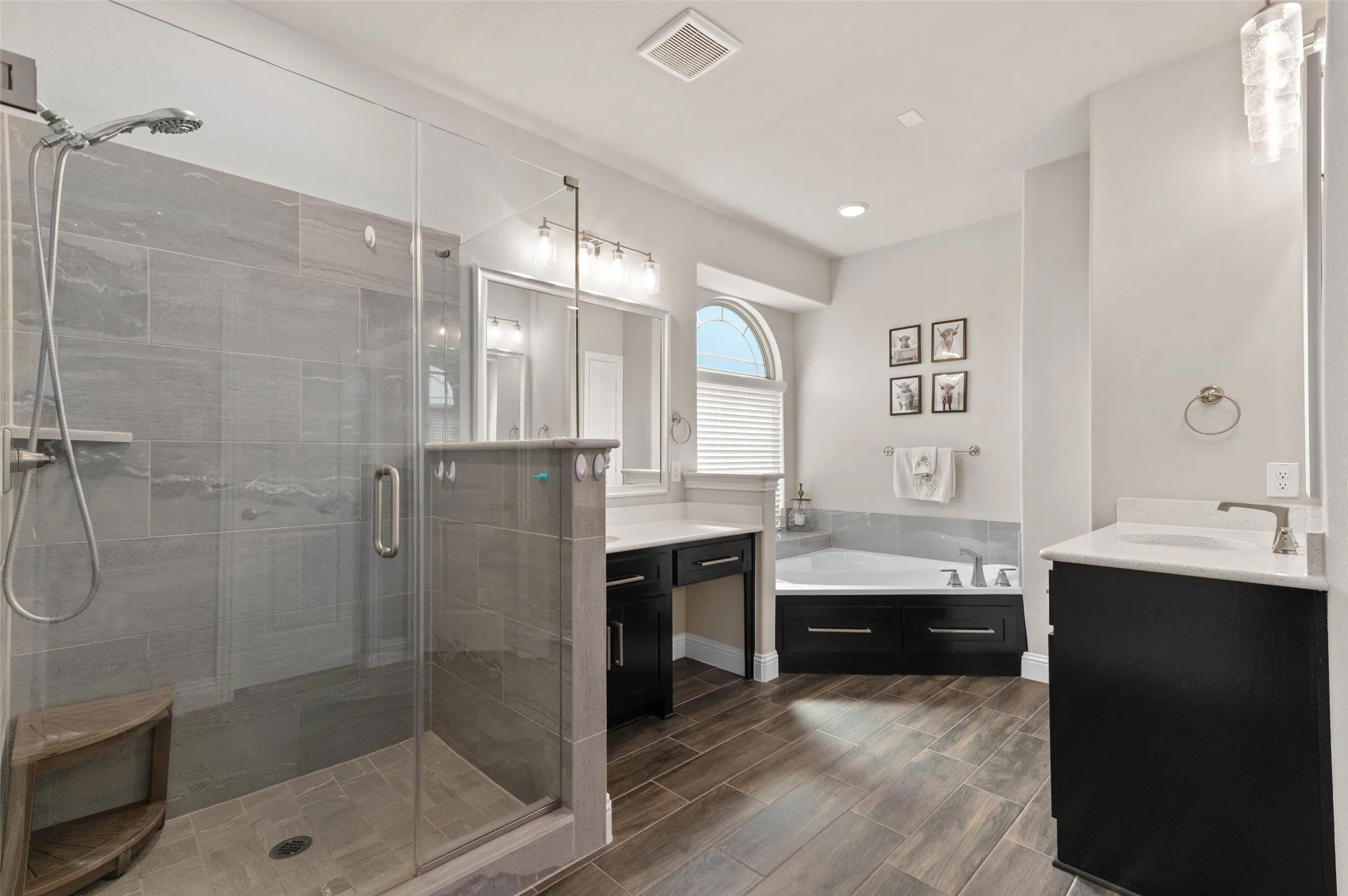 Spacious Primary bathroom featuring a glass-enclosed shower with large format tile, wood-finish flooring, a soaking tub with tile surround, and dual vanity areas with dark cabinetry