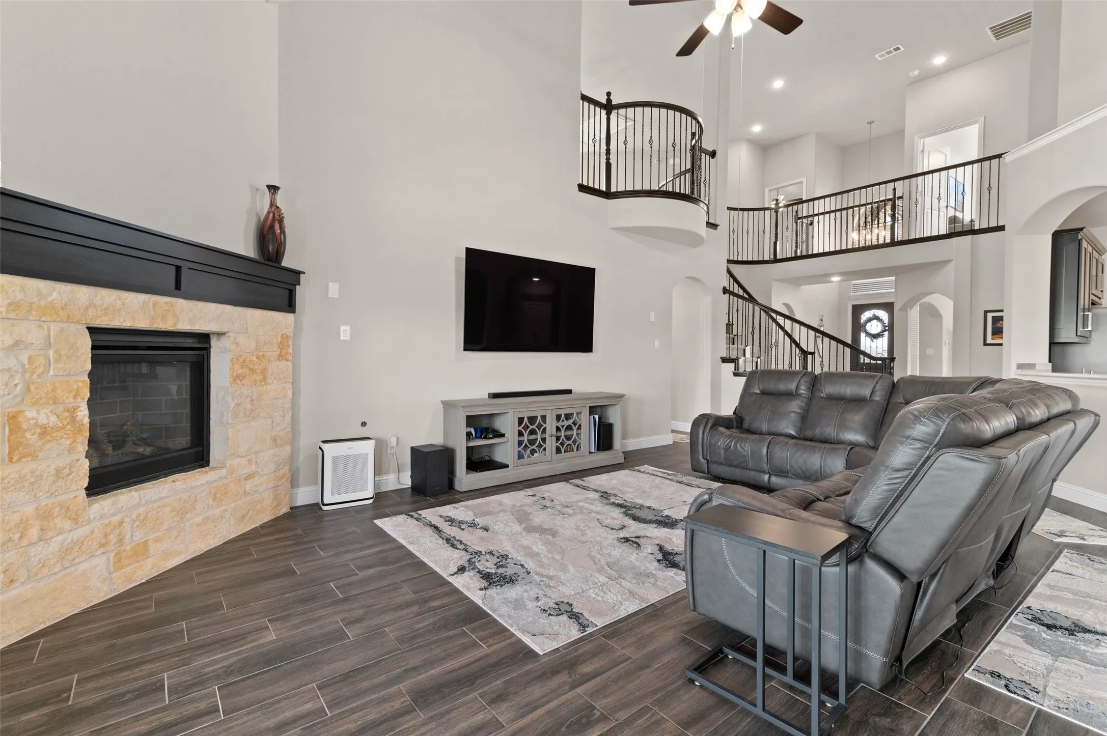 Two-story living area featuring a stone fireplace with a dark wood mantle, wood-look tile flooring, and a decorative wrought iron staircase and balcony