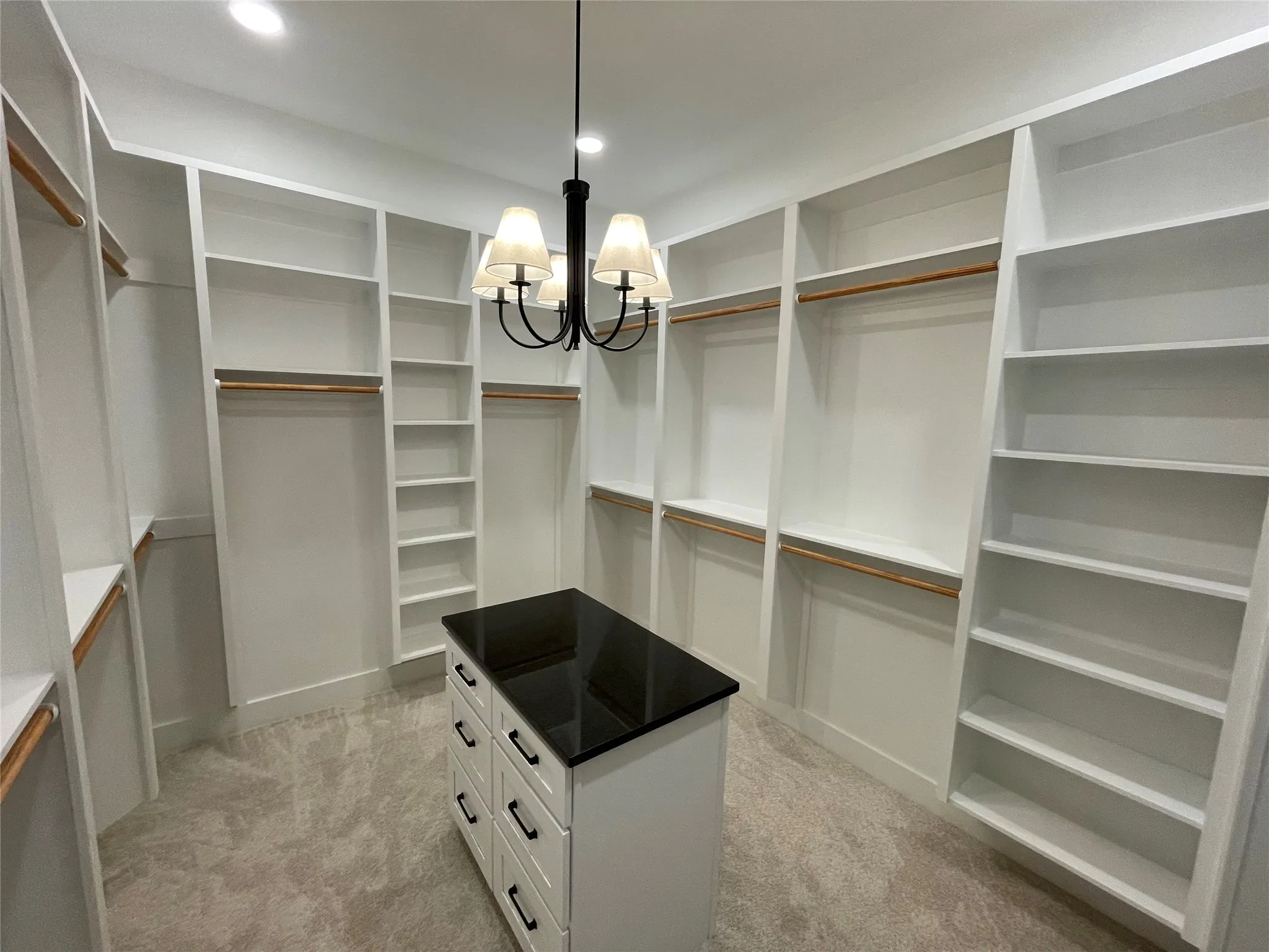 Walk-in closet featuring custom white built-in shelving, hanging rods, an island with a dark countertop and drawers, recessed lighting, and a central chandelier