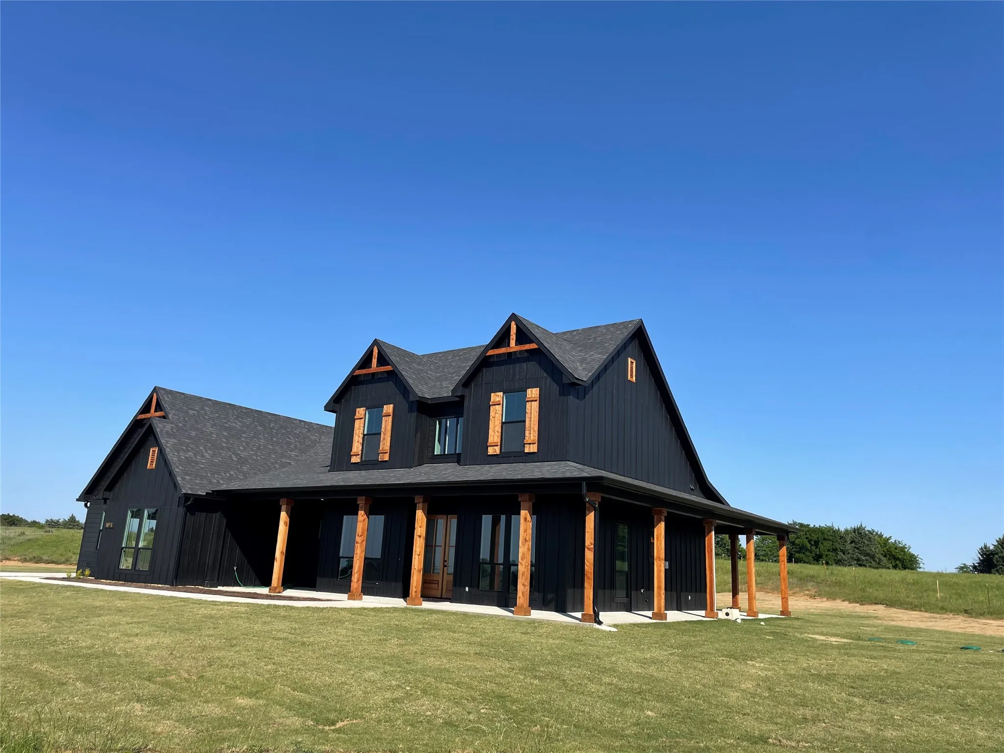Modern farmhouse exterior featuring dark board and batten siding, a covered wraparound porch with wood columns, dark shingle roofing, and decorative wood shutters
