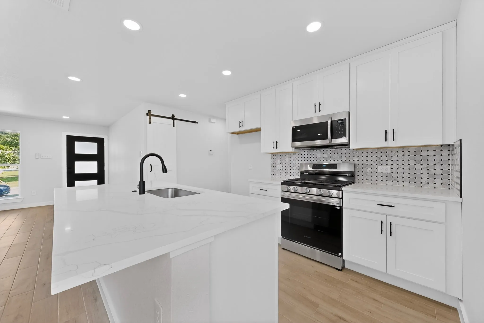 Modern kitchen featuring an expansive island with an undermount sink and matte black faucet, stainless steel appliances, white cabinetry with contrasting hardware, and a graphic tile backsplash