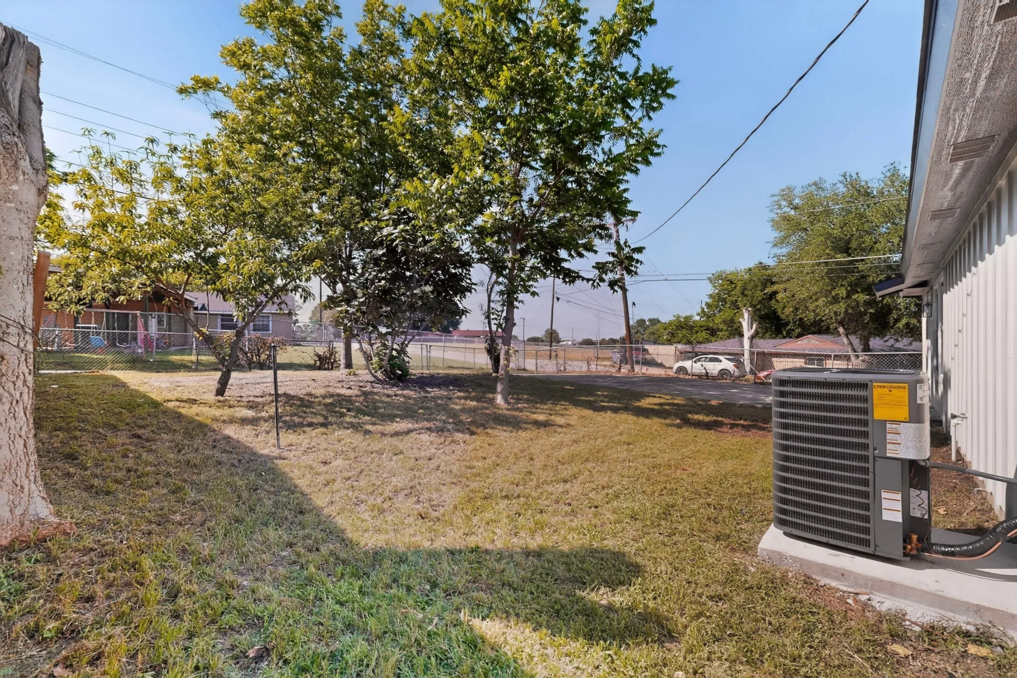 Expansive grassy area with mature trees and a chain-link fence, featuring an exterior air conditioning unit on a concrete pad