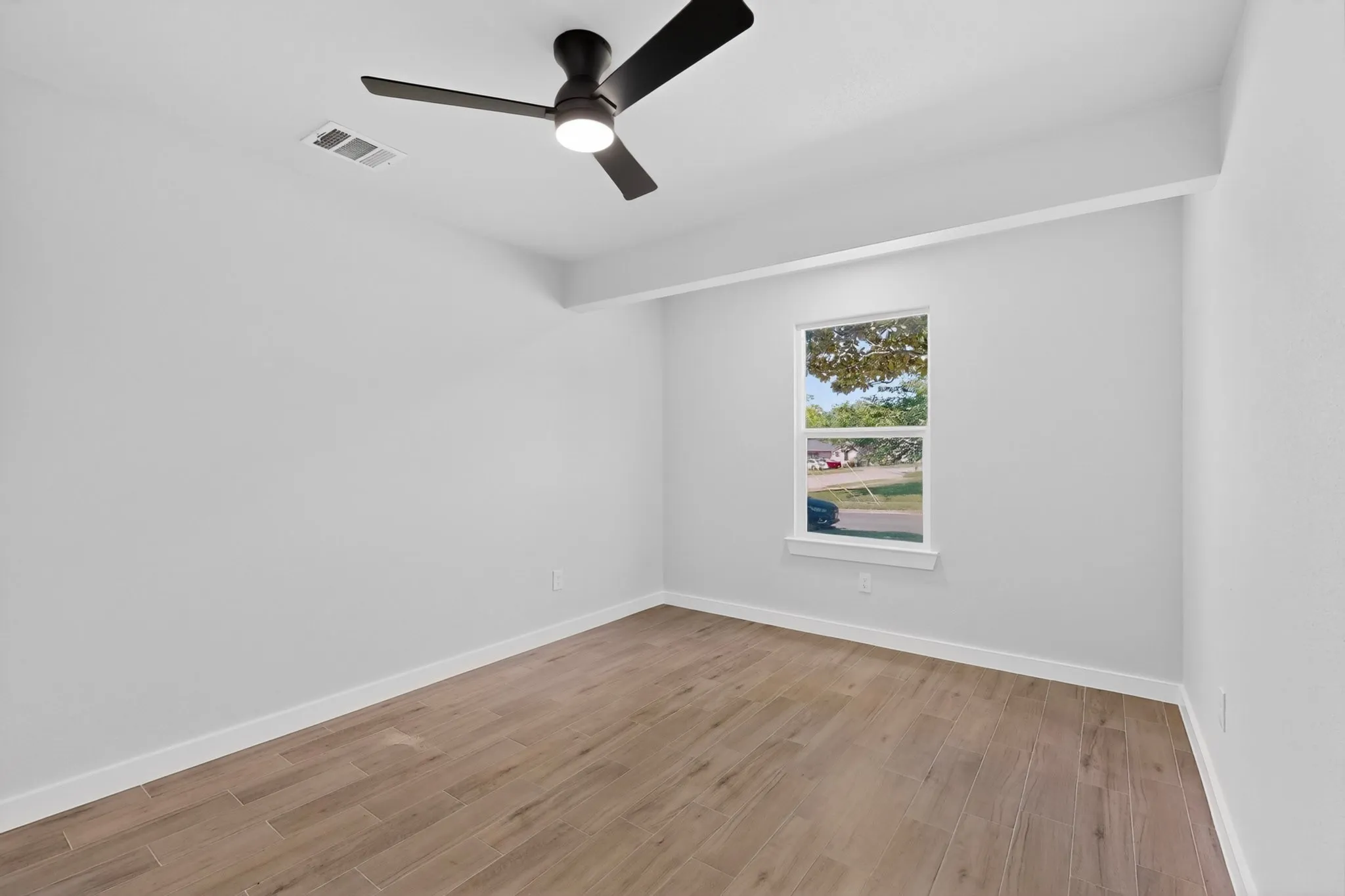 Spacious room featuring wood-finish flooring, a modern black ceiling fan with integrated lighting, a single window, and crisp white walls