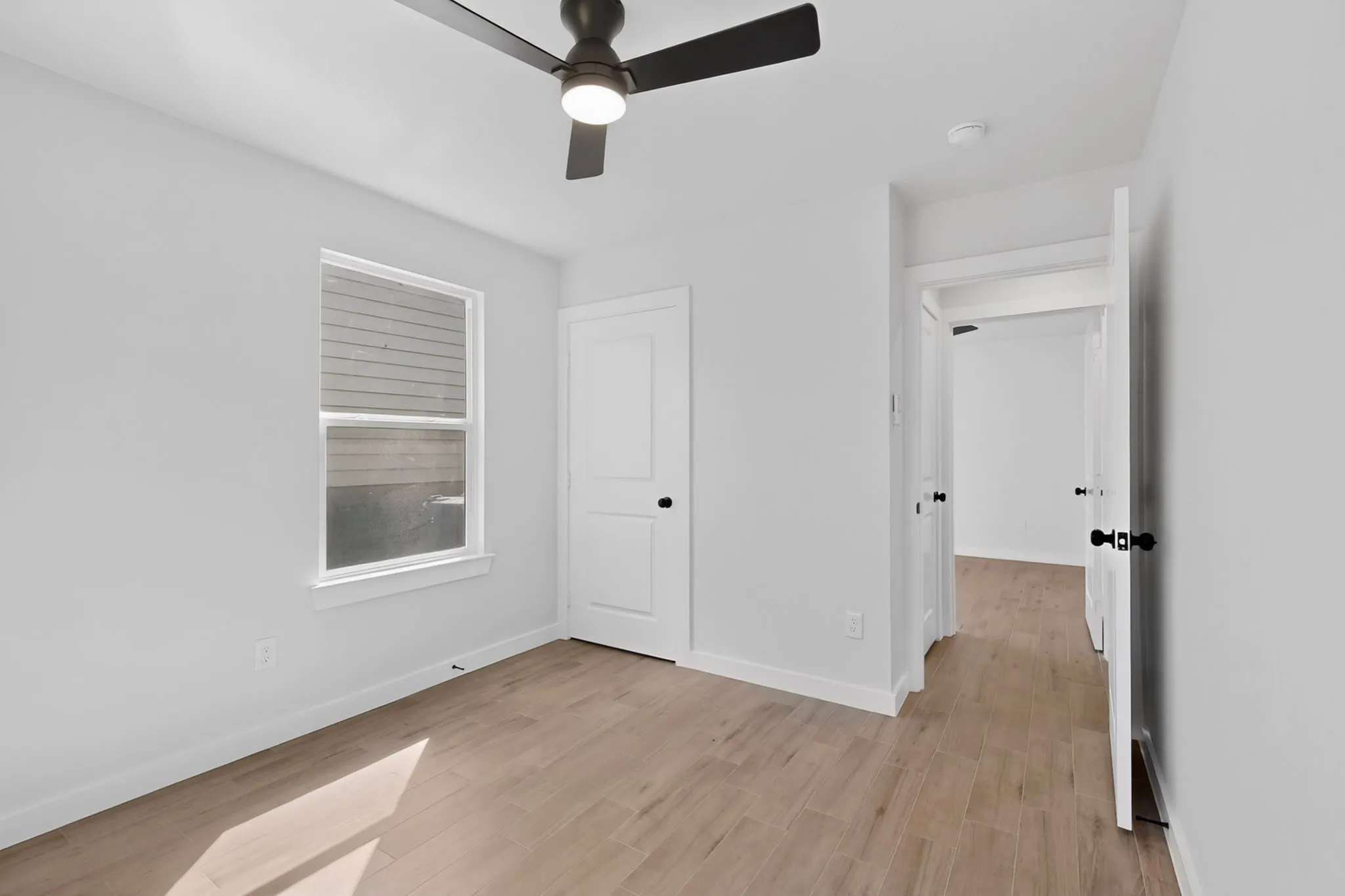 Room featuring light wood-finish flooring, white walls, and a modern black ceiling fan with integrated lighting