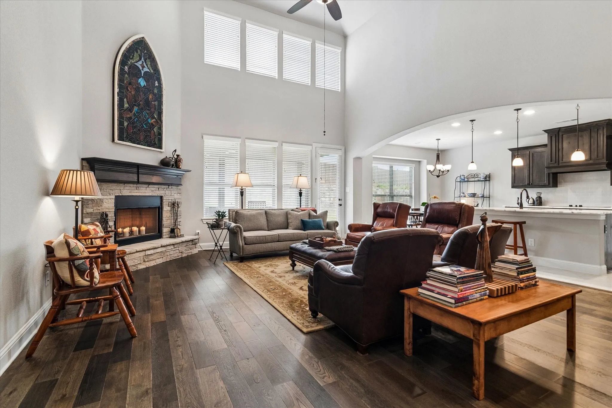 Spacious living area featuring high ceilings, wood-finish flooring, a stone-surround fireplace with a dark wood mantle, and an open archway to the kitchen
