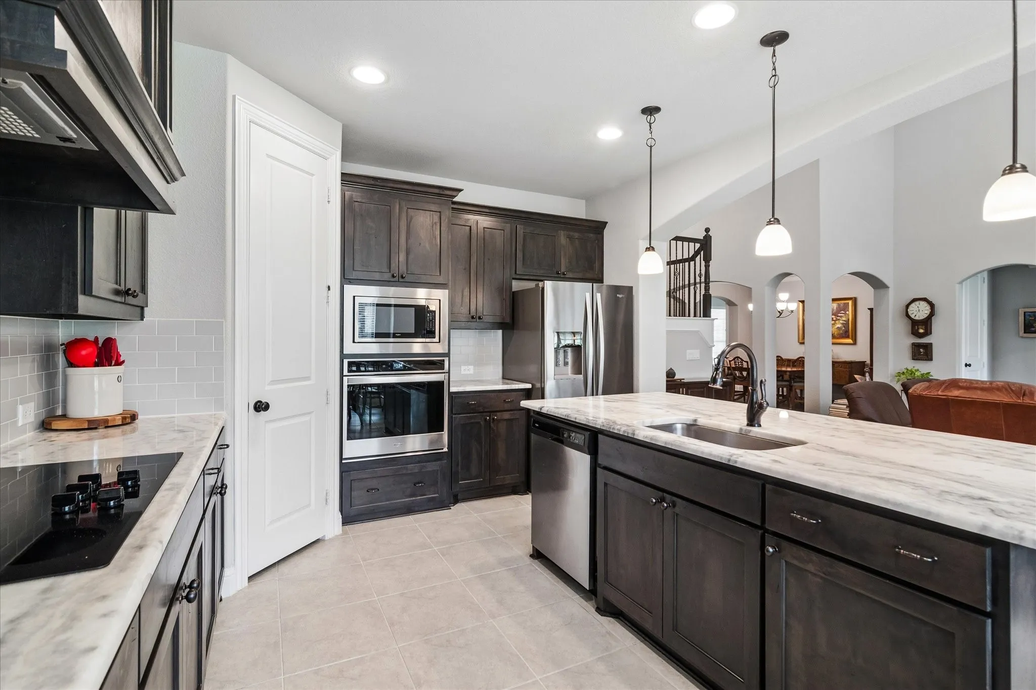 Modern kitchen featuring a large island with an undermount sink, stainless steel appliances, dark wood cabinetry, light marble-finish countertops, and a subway tile backsplash