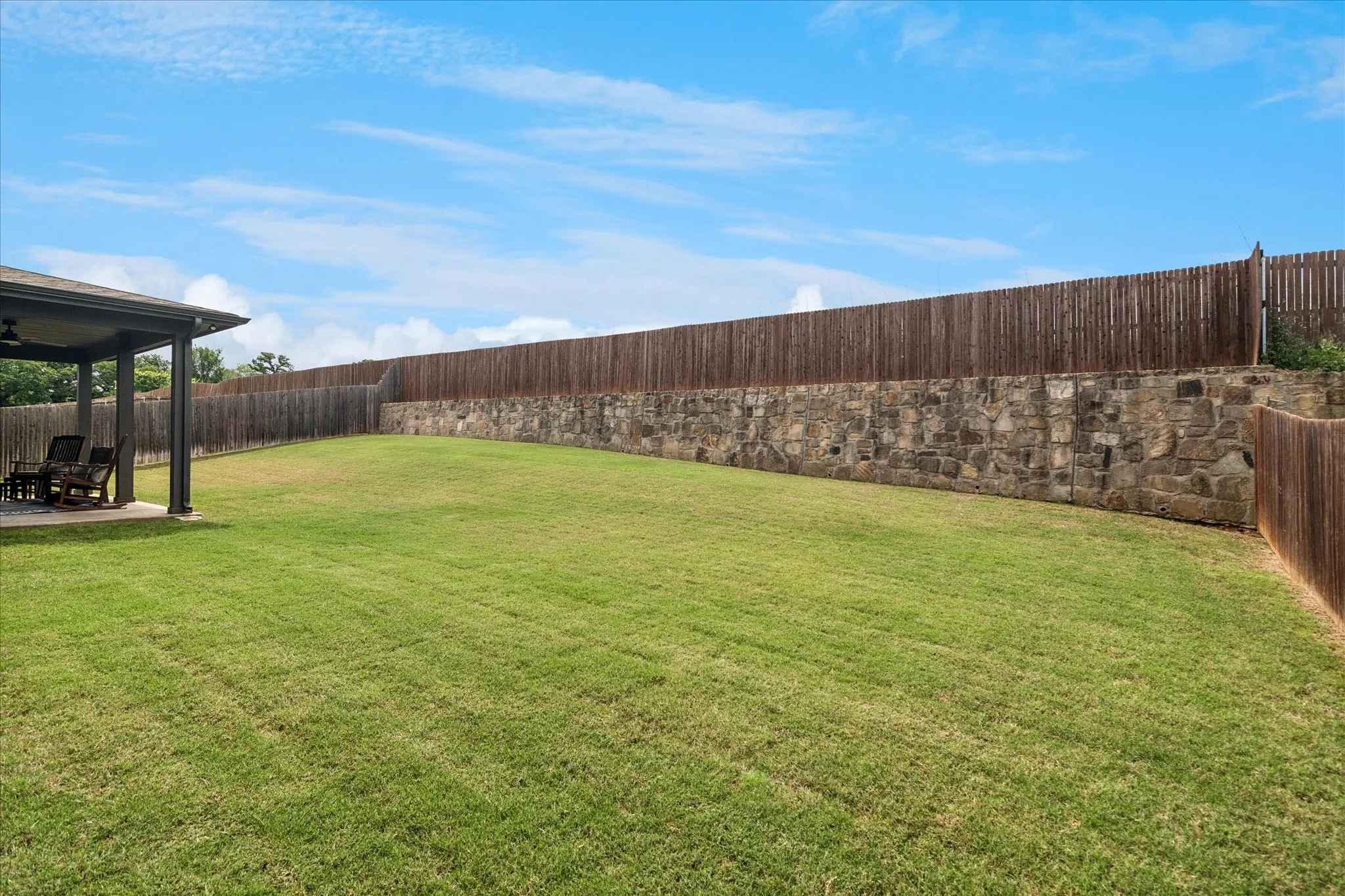Expansive backyard featuring a stone retaining wall, wood privacy fencing, and a covered patio with ceiling fan