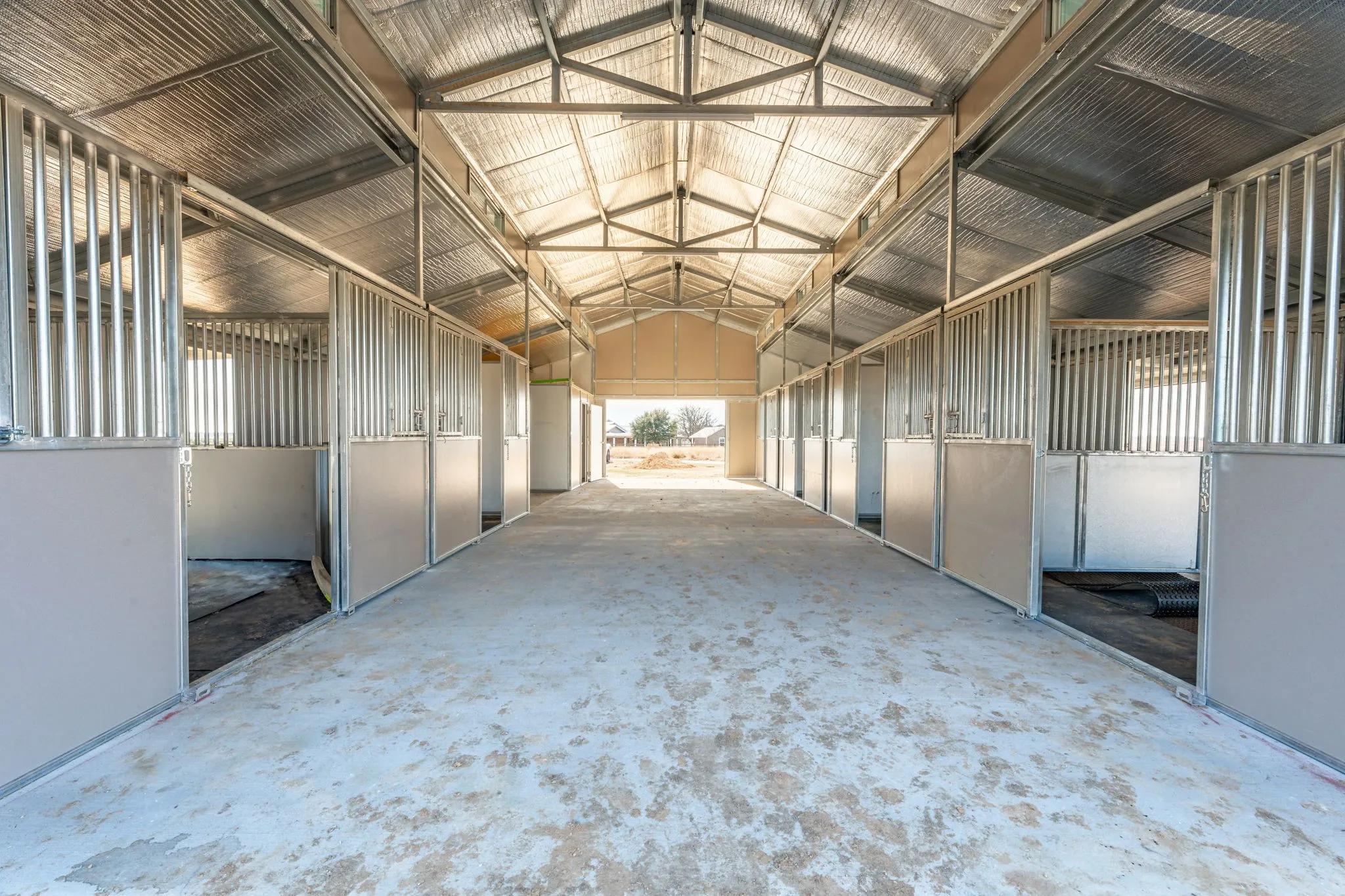 Interior barn aisle featuring a concrete floor, multiple stalls with metal grates and solid lower panels, exposed metal truss ceiling, and a corrugated metal roof