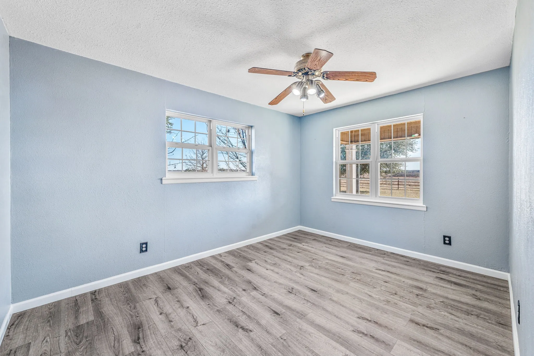 Spacious room featuring light blue walls, wood-finish flooring, two large windows, a ceiling fan with integrated lighting, and white baseboards