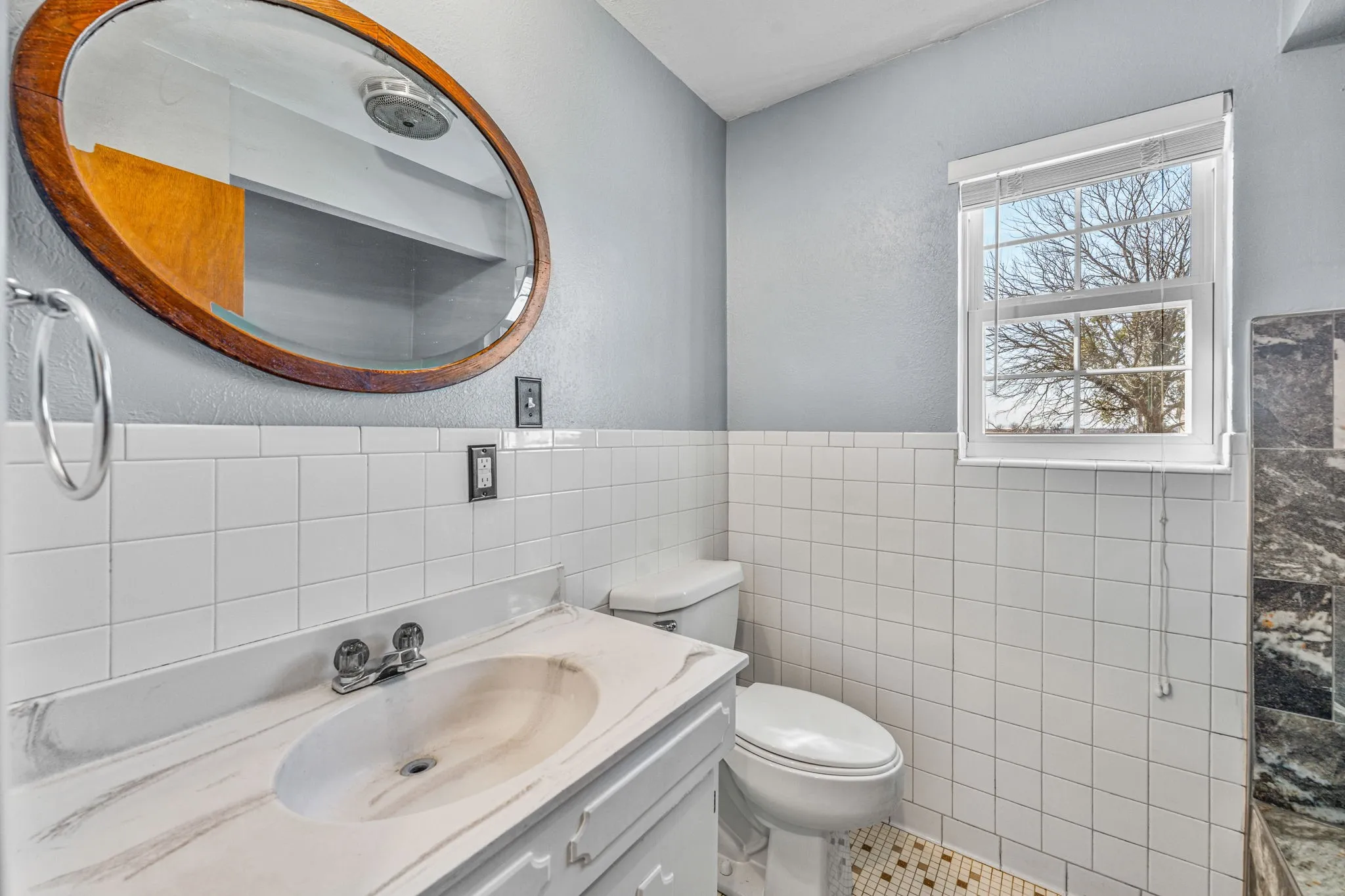 Bathroom featuring a white tile wainscoting, a white vanity with an integrated sink and light-colored countertop, and an oval wood-framed mirror