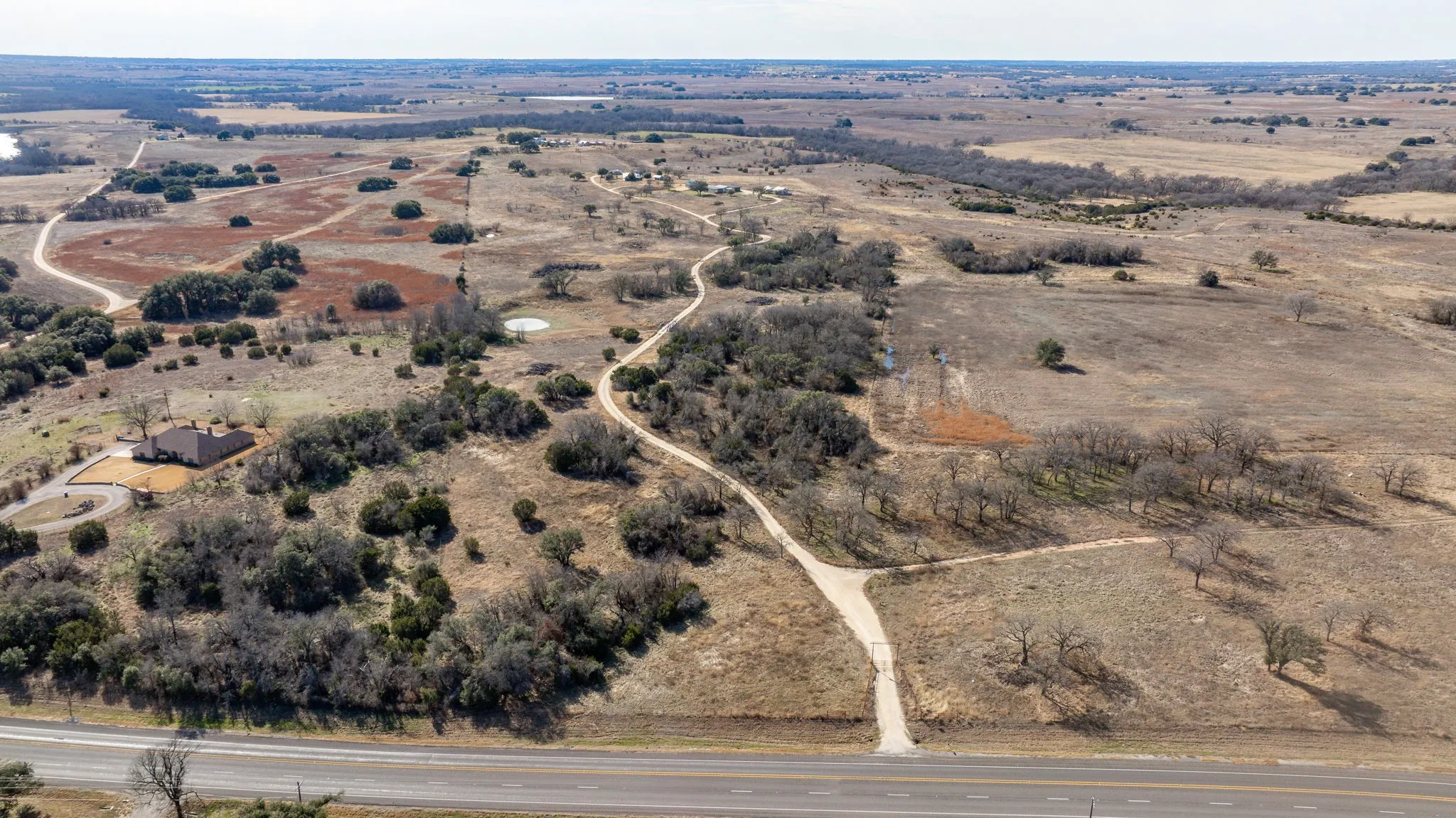 Expansive rural property featuring a single-story dwelling with a brown shingle roof, surrounded by varied terrain including open fields and wooded areas