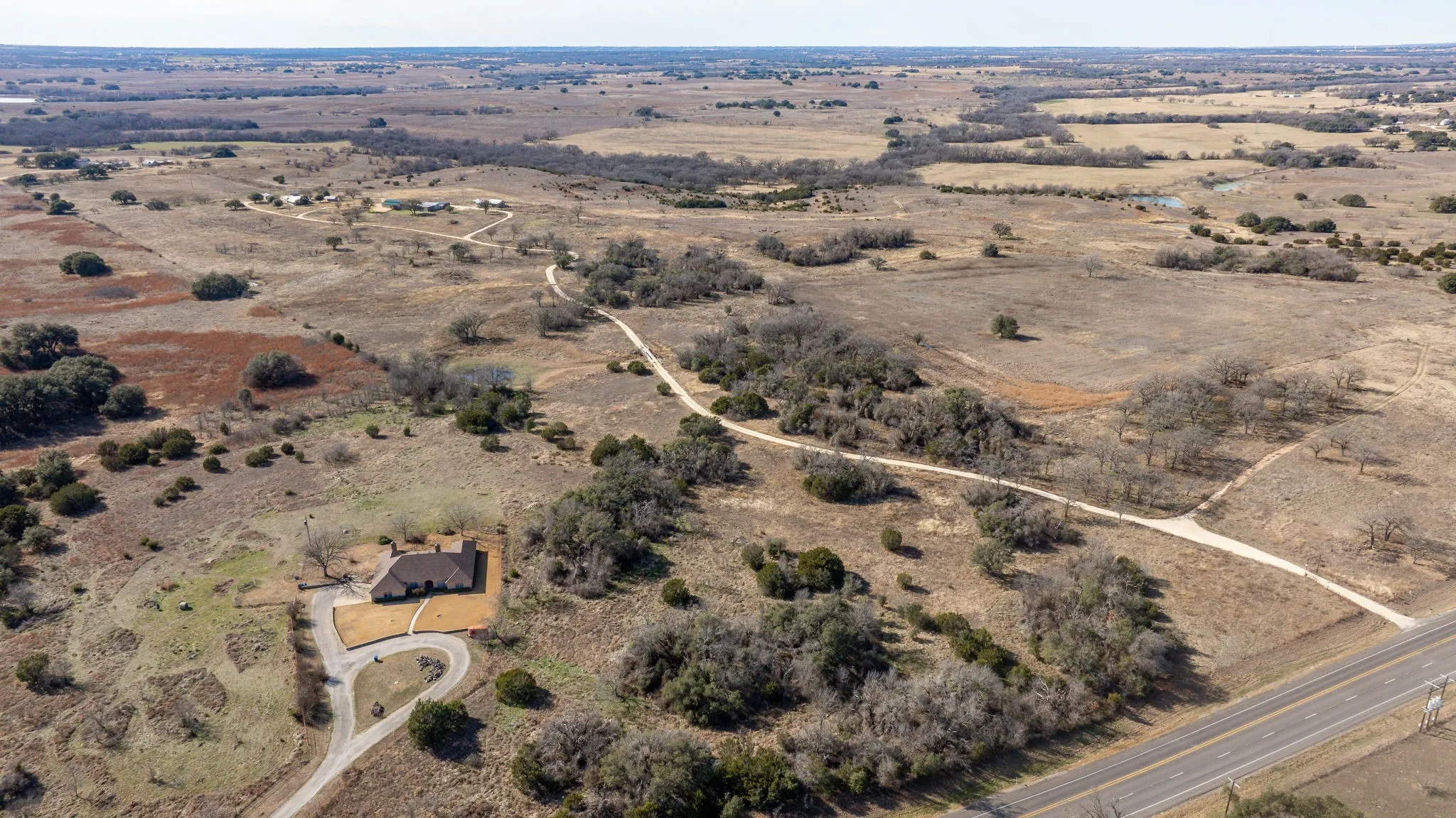 Expansive rural property featuring a brown-toned residence with a paved circular driveway, multiple unpaved paths, scattered mature trees, and a distant road