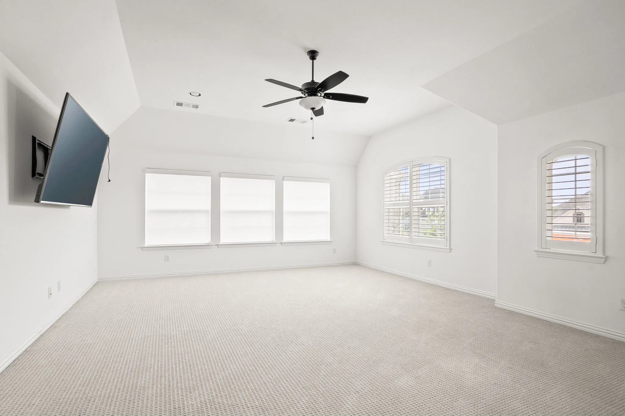 Spacious room featuring vaulted ceilings, light-colored carpet flooring, multiple windows with roller shades, and two arched windows with plantation shutters