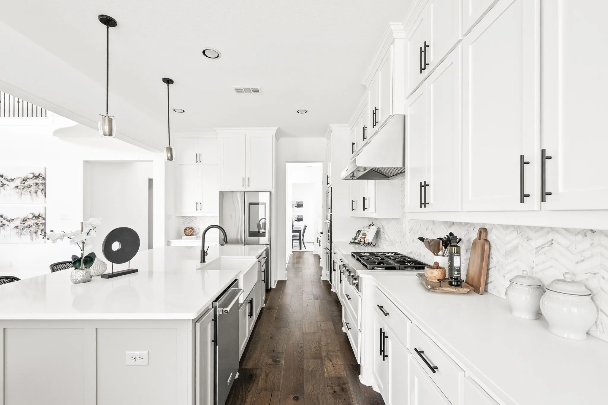 Modern kitchen featuring an expansive island with a built-in sink and pendant lighting, white shaker-style cabinetry with matte black hardware, stainless steel appliances, and wood-finish flooring