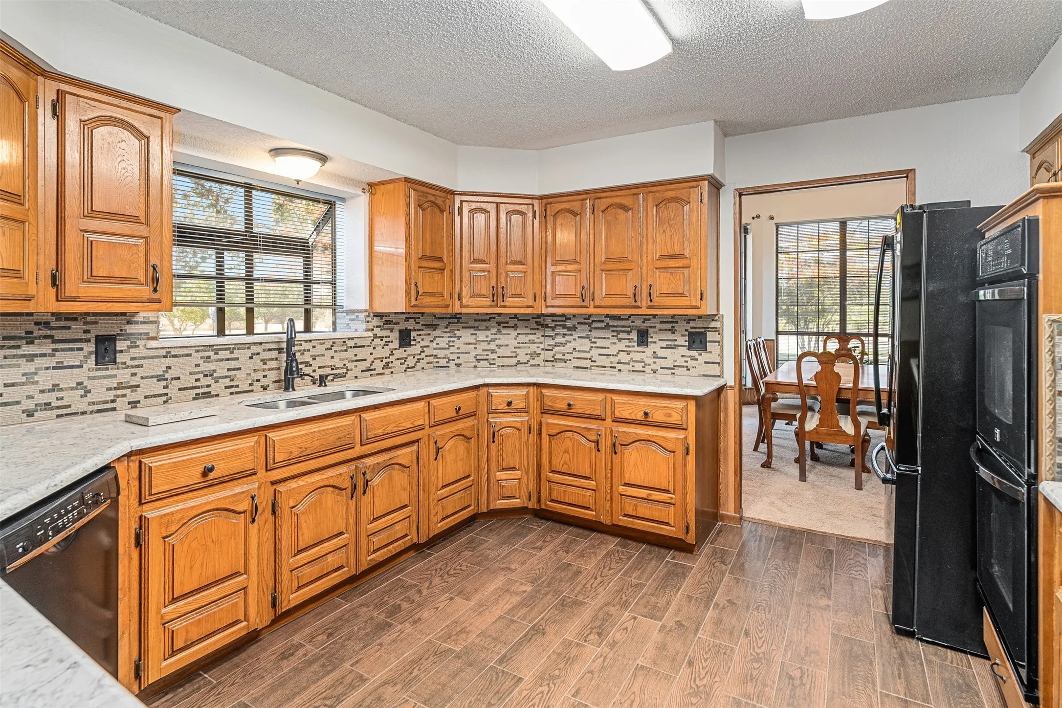 Kitchen with a textured ceiling, decorative backsplash, brown cabinetry, black appliances, and healthy amount of natural light