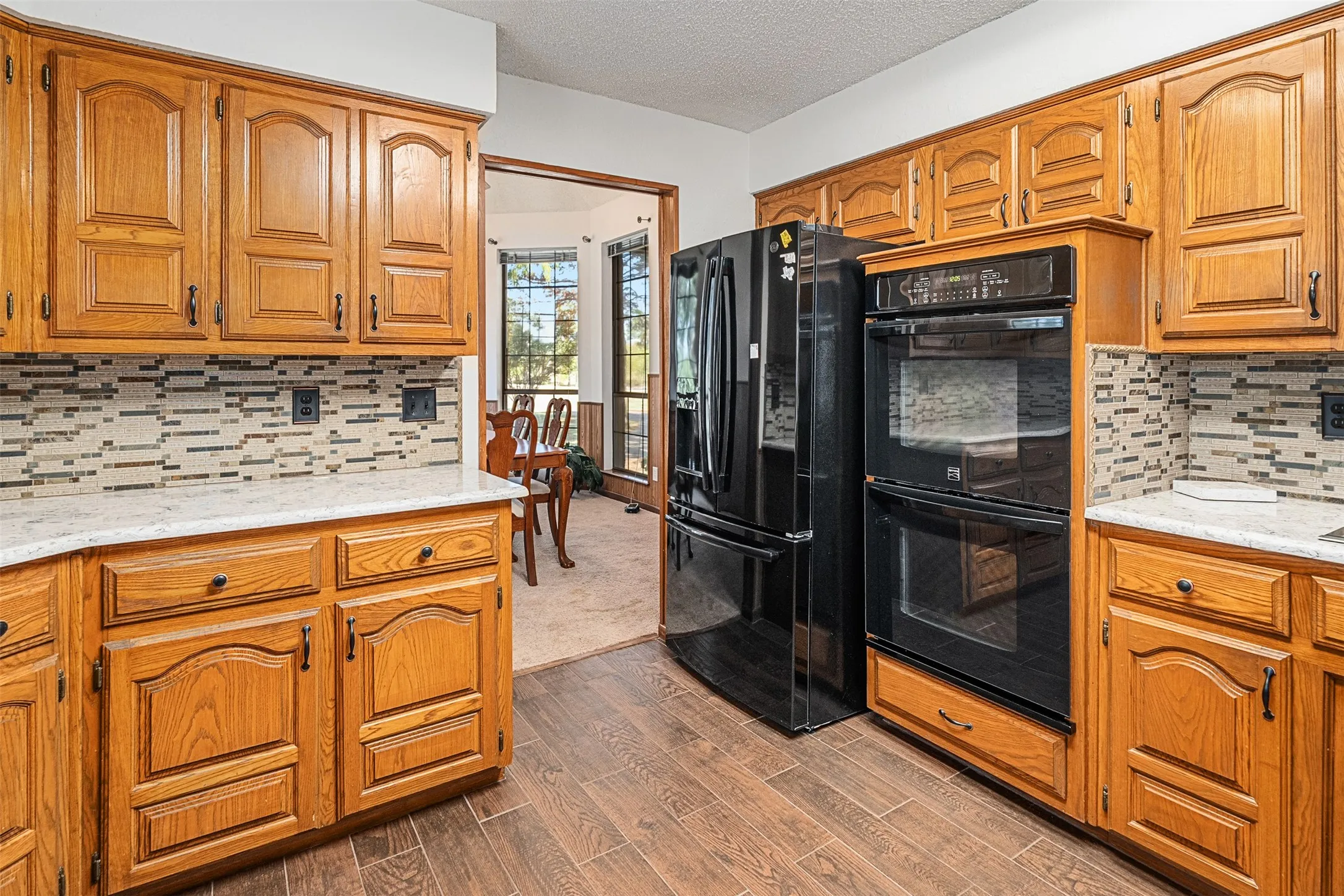 Kitchen with backsplash, black appliances, a textured ceiling, brown cabinets, and dark wood-style floors