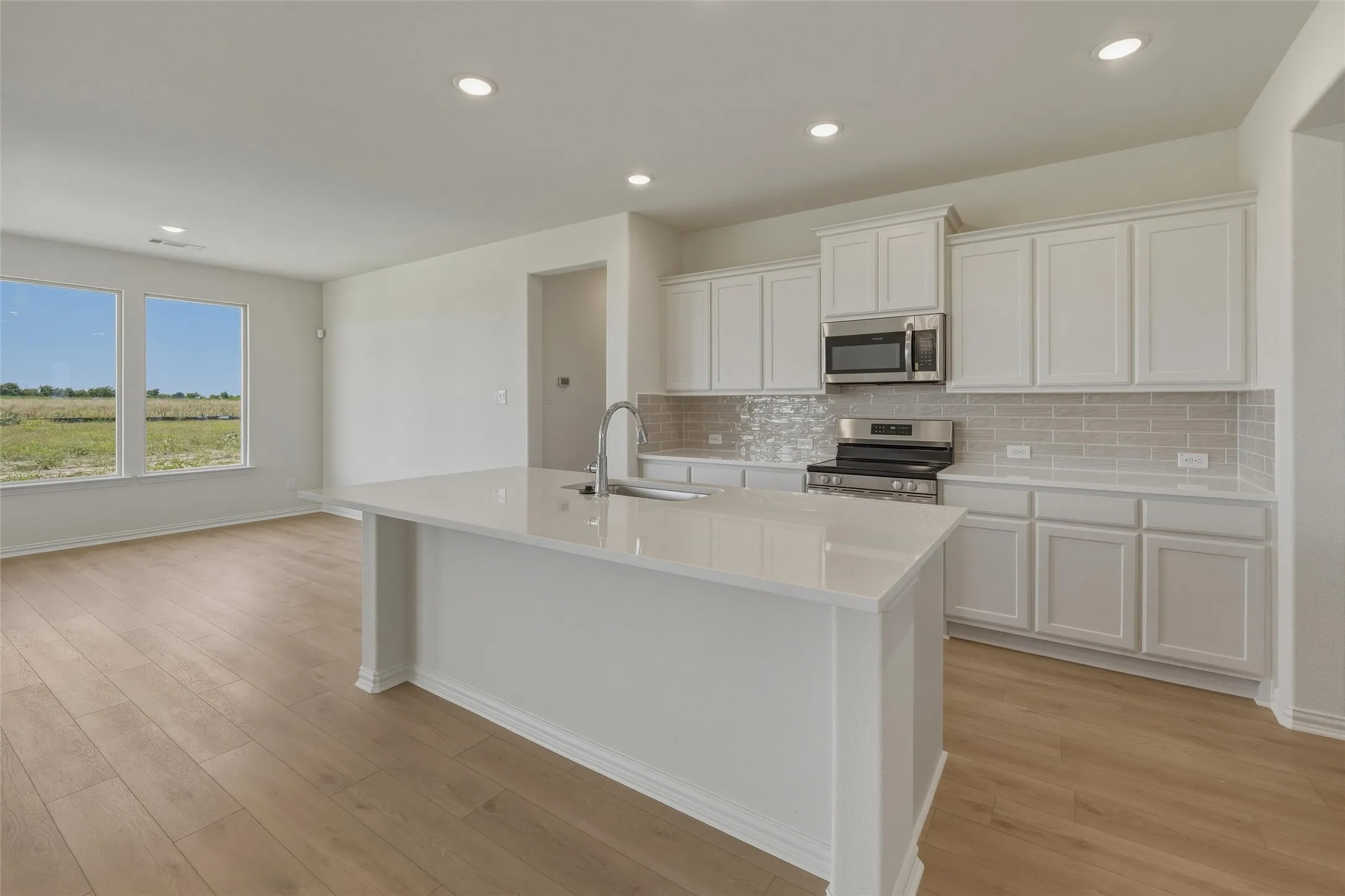 Kitchen island with a built-in sink and stainless steel faucet, white cabinetry, integrated stainless steel microwave, gas range, light-toned subway tile backsplash, and recessed lighting