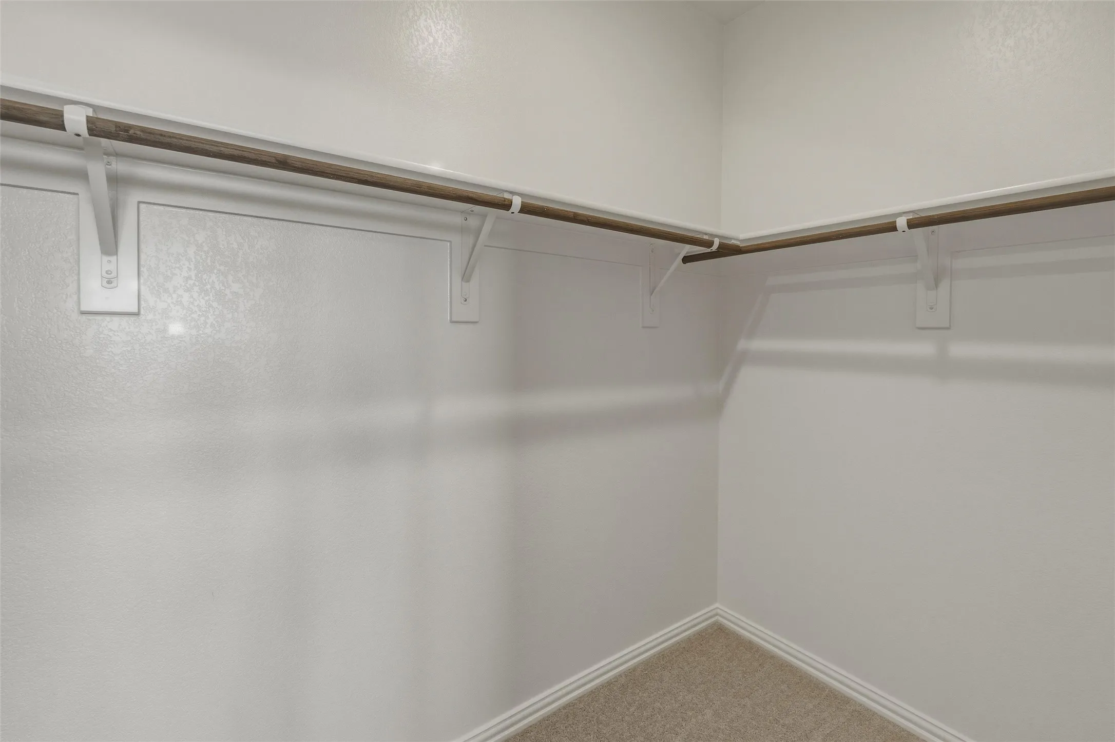 Walk-in closet featuring white walls, wood-finish hanging rods, white support brackets, and light-colored carpet