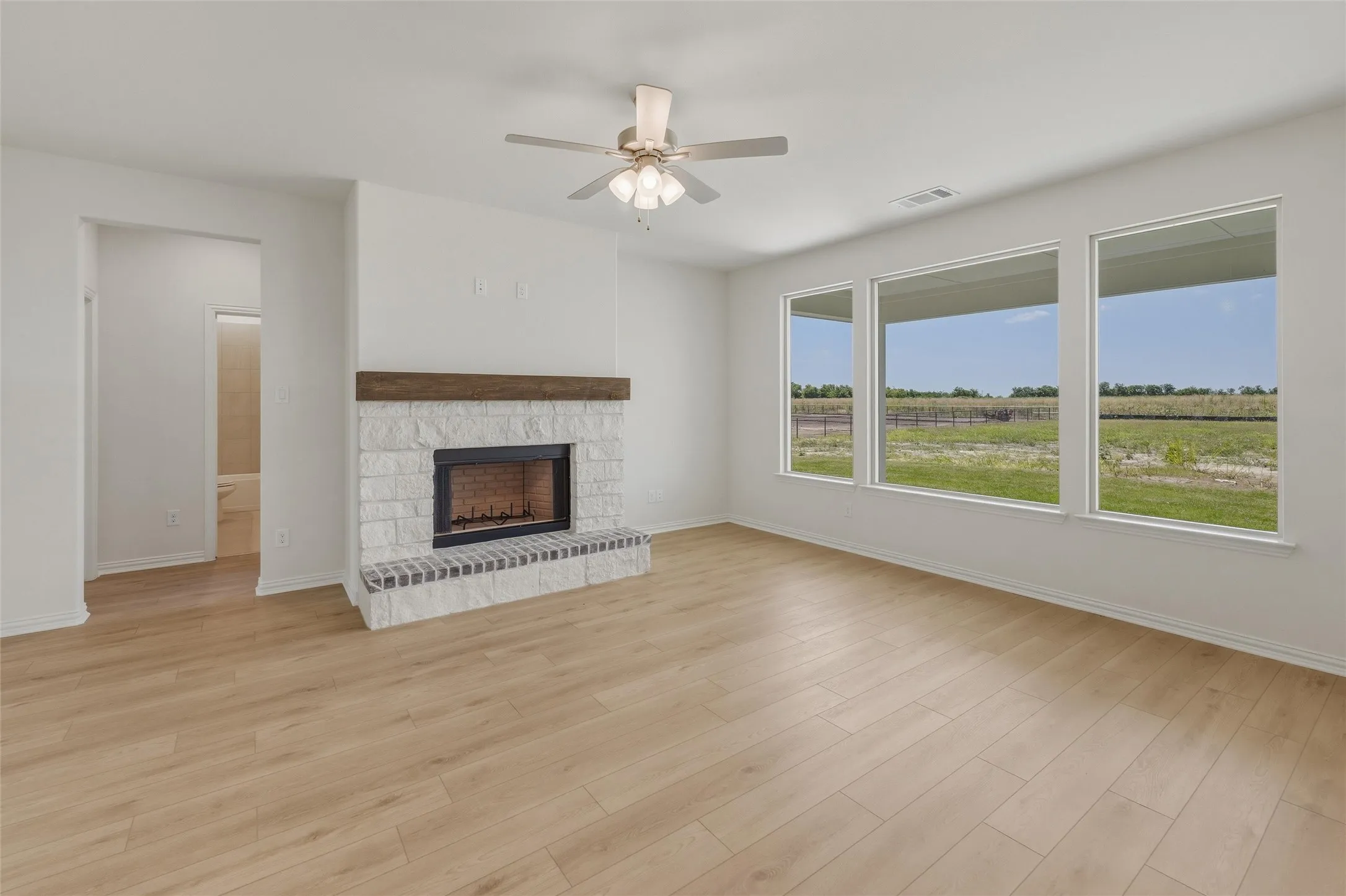 Living space featuring a whitewashed brick fireplace with a wood-finish mantel, light wood-finish flooring, and a ceiling fan with integrated lighting
