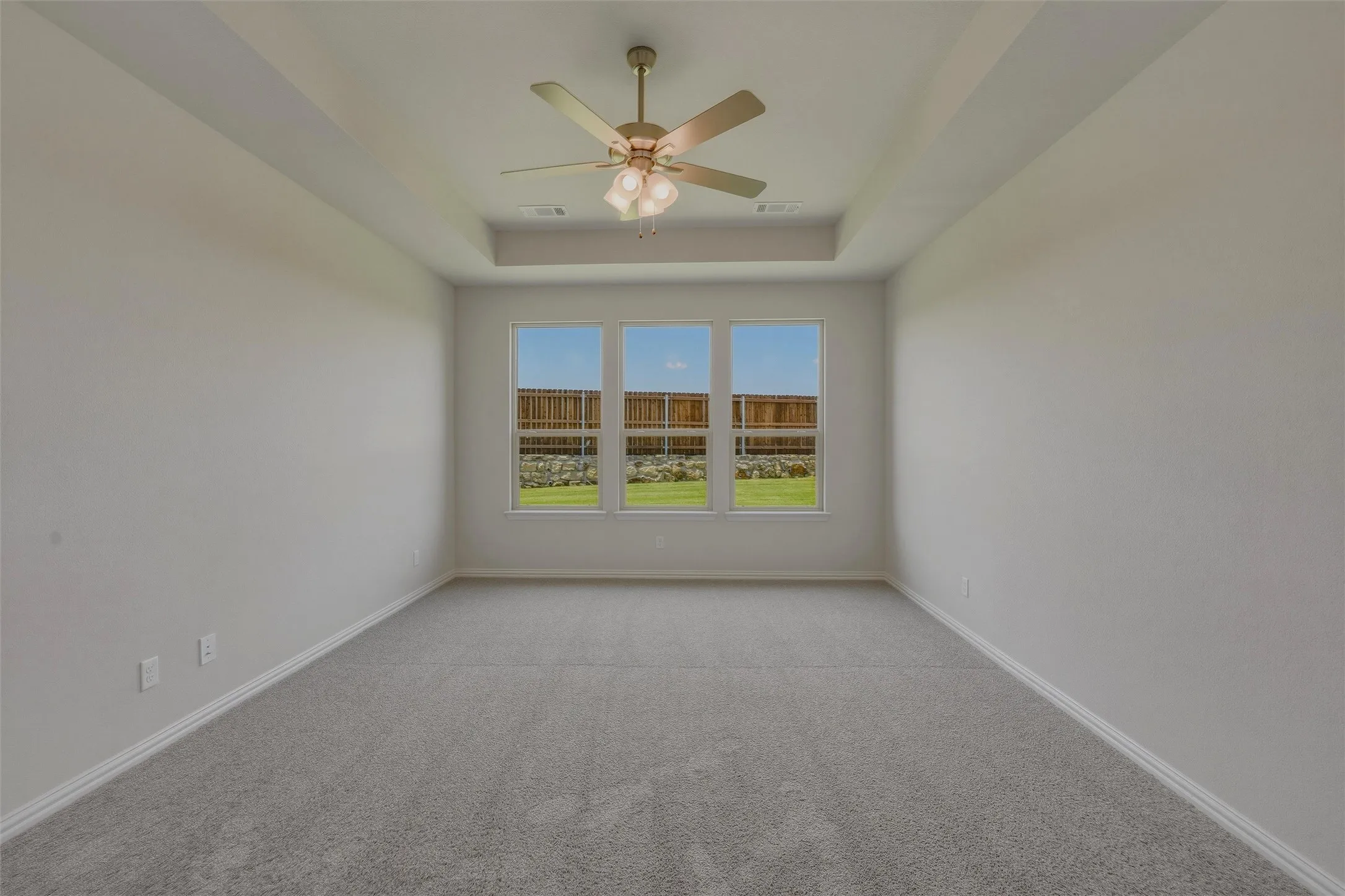 Spacious room featuring a tray ceiling with a ceiling fan, three large windows, gray carpeting, and white baseboards