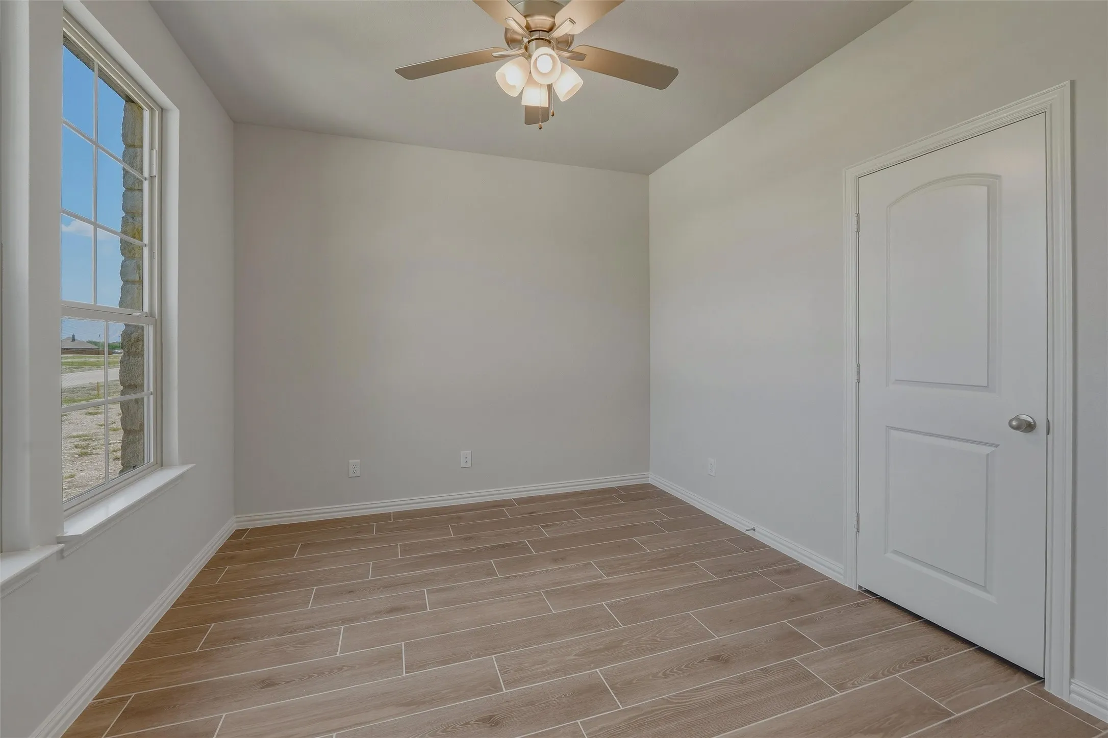 Interior room featuring a large double-hung window, light wood-finish flooring, a ceiling fan with integrated lighting, and a paneled interior door