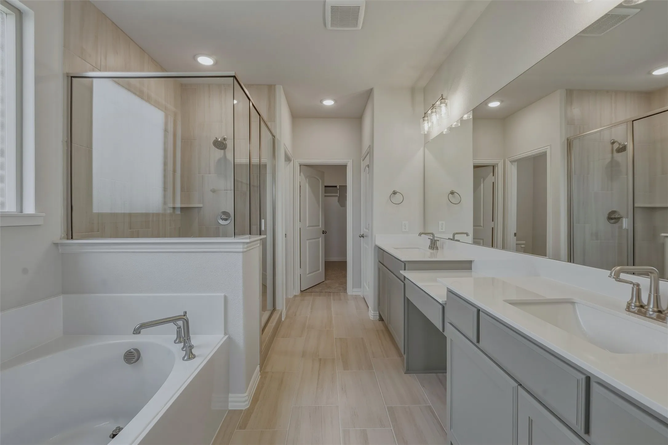 Spacious bathroom featuring a built-in soaking tub, a glass-enclosed shower with tile surround, a double vanity with undermount sinks, and light wood-finish flooring