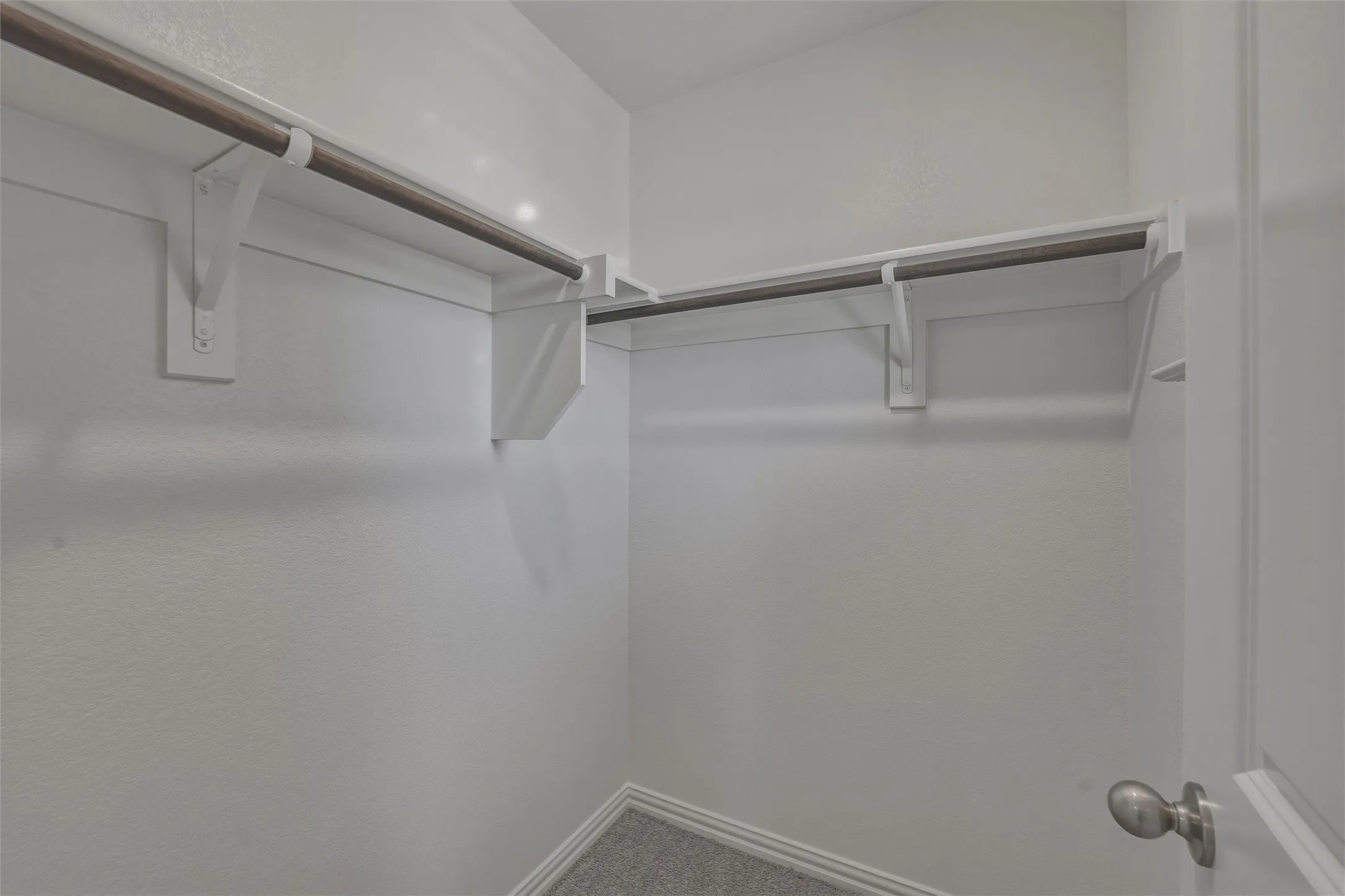 Walk-in closet featuring white walls, gray carpet flooring, and two metal hanging rods with white supports