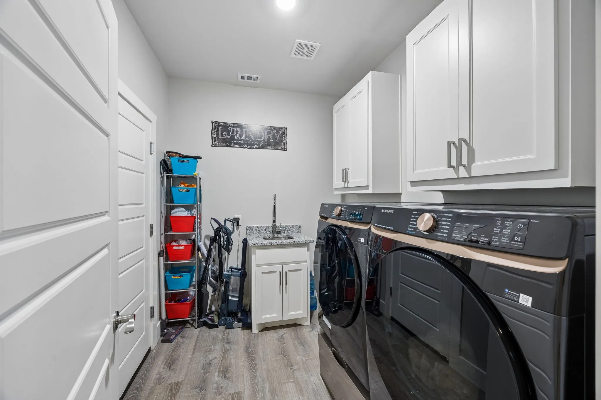 Dedicated laundry area featuring wood-finish flooring, white cabinetry with silver hardware, and a utility sink with a granite-look countertop