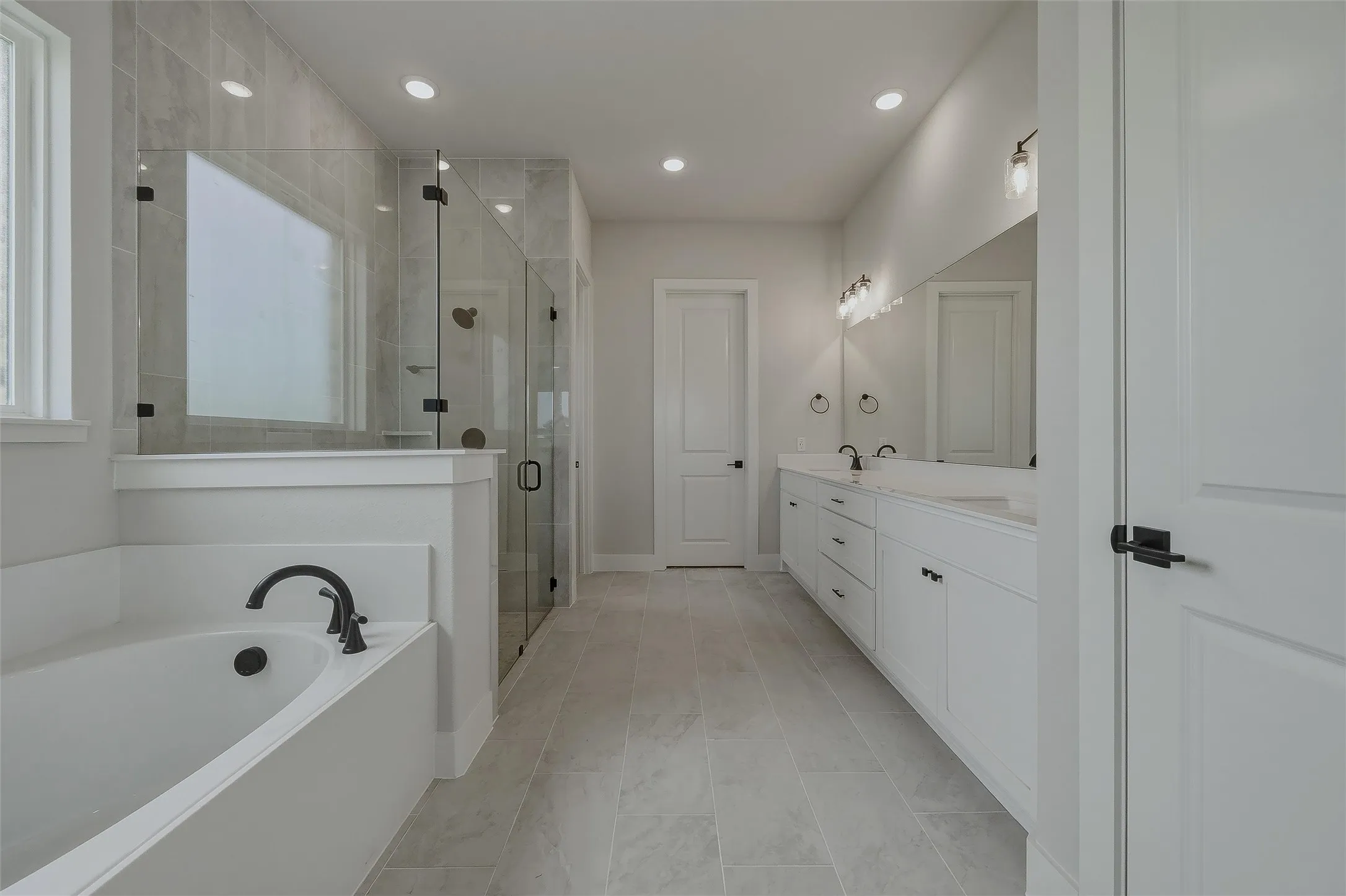Freestanding soaking tub with matte black fixtures, glass-enclosed shower with tile surround, dual vanity with undermount sinks, and tile flooring