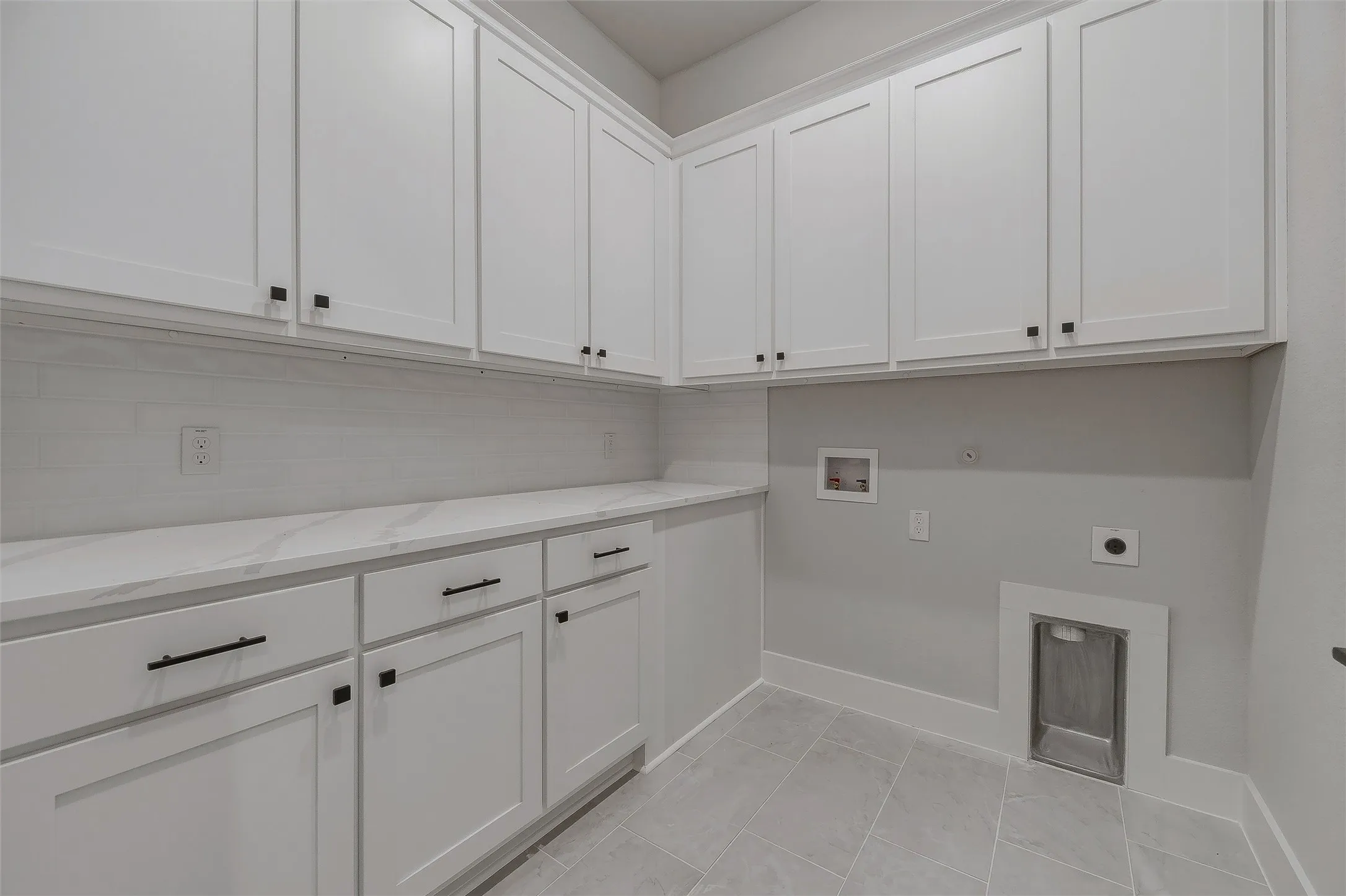 Extensive white shaker cabinetry, white countertop, white subway tile backsplash, contemporary black hardware, and gray tile flooring