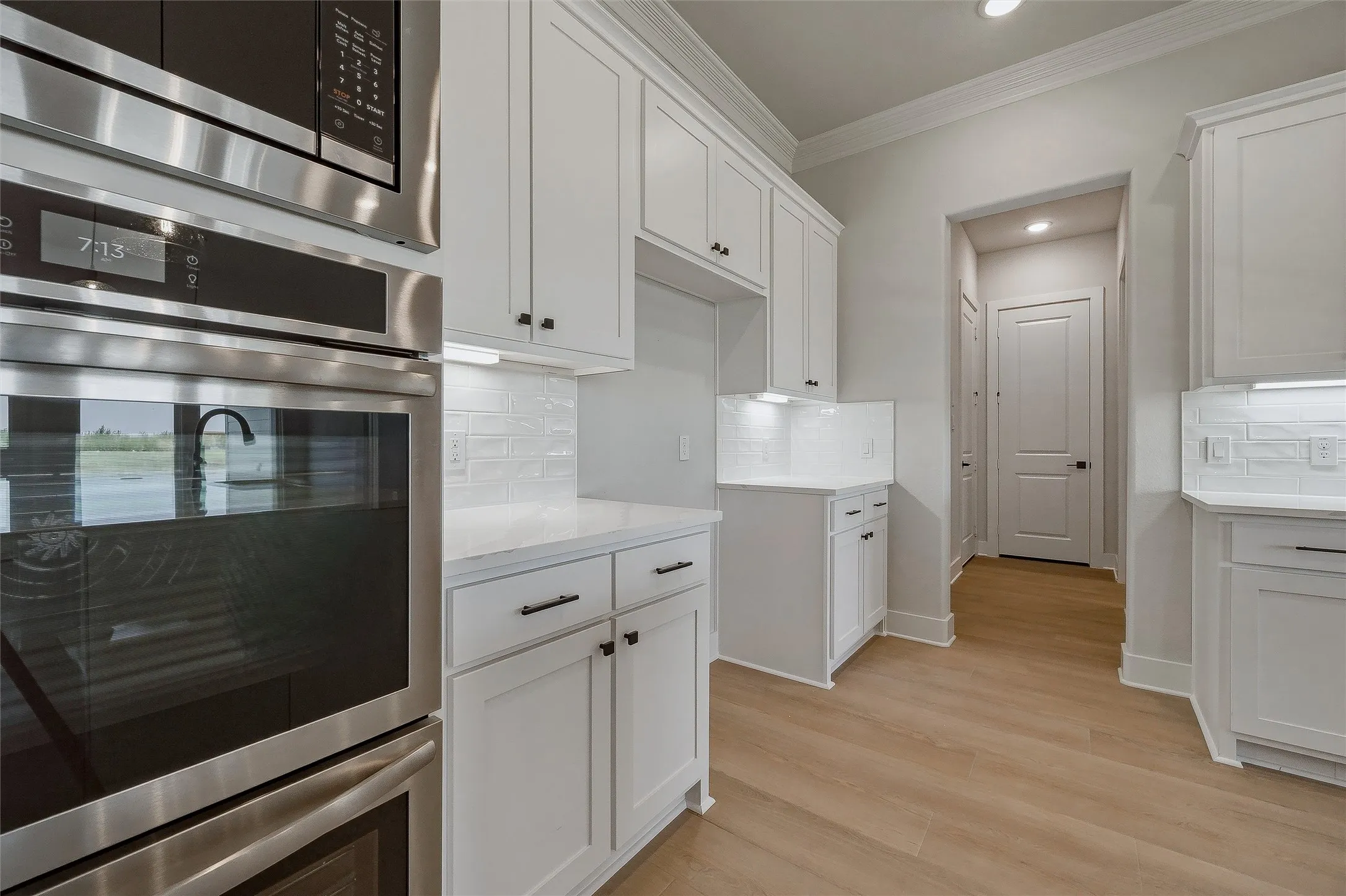 Stainless steel built-in oven and microwave, white shaker-style cabinetry, light-toned wood-finish flooring, white subway tile backsplash, and white countertops