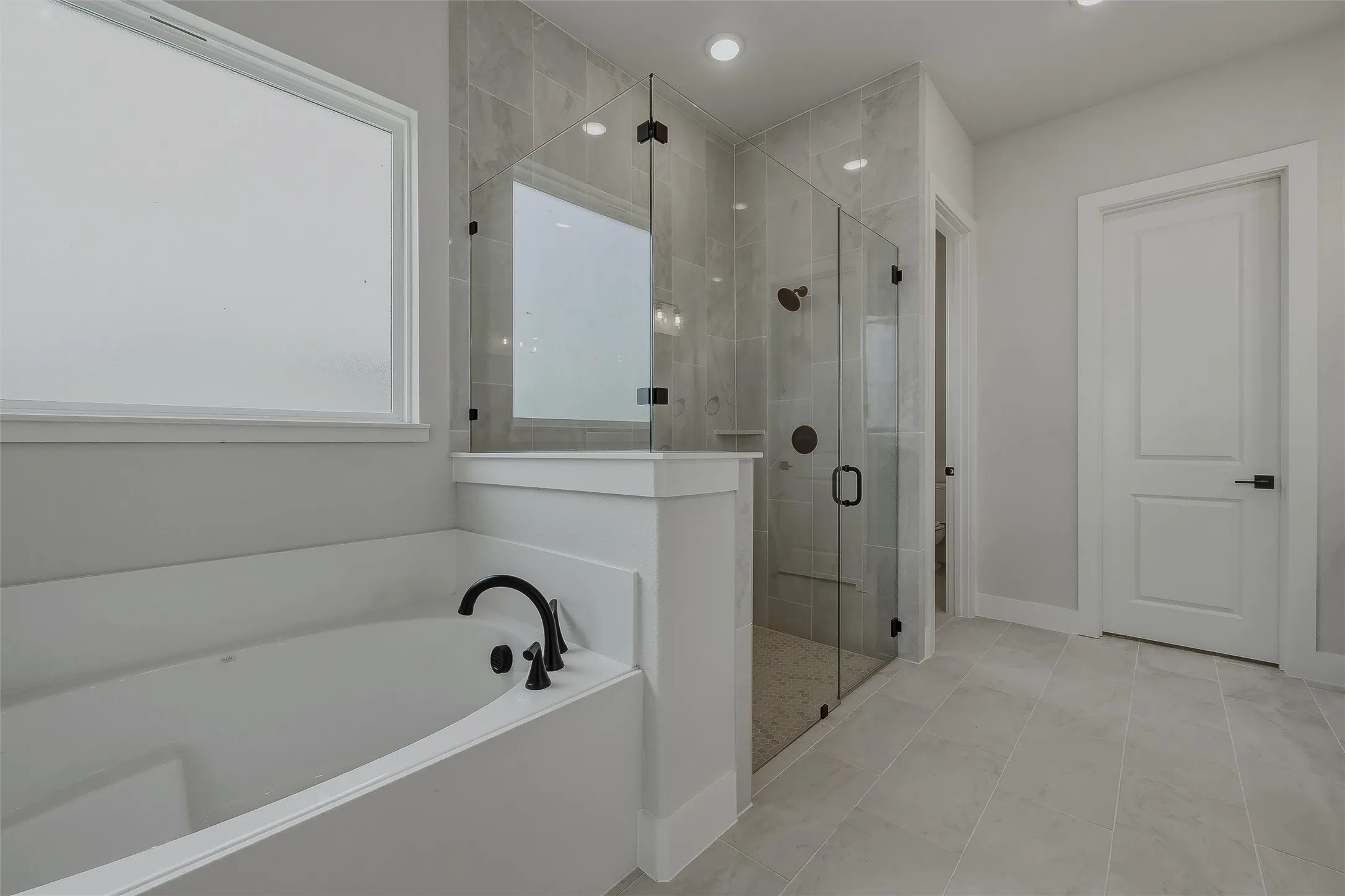 Freestanding soaking tub with matte black fixtures, glass-enclosed shower with tile surround, and large format tile flooring