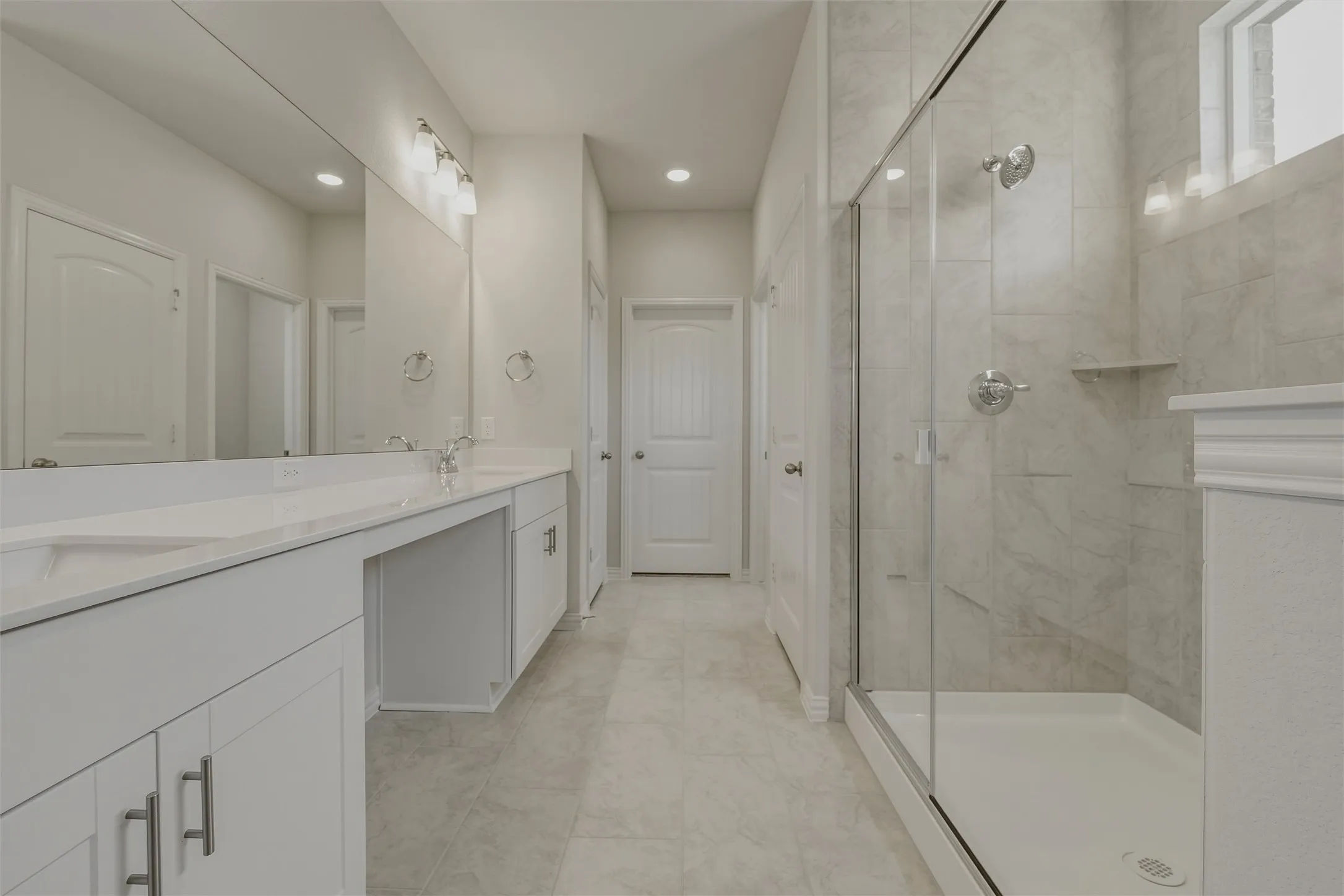 Spacious bathroom featuring a dual vanity with white cabinetry, a large glass-enclosed shower, polished chrome fixtures, and neutral-toned tile flooring