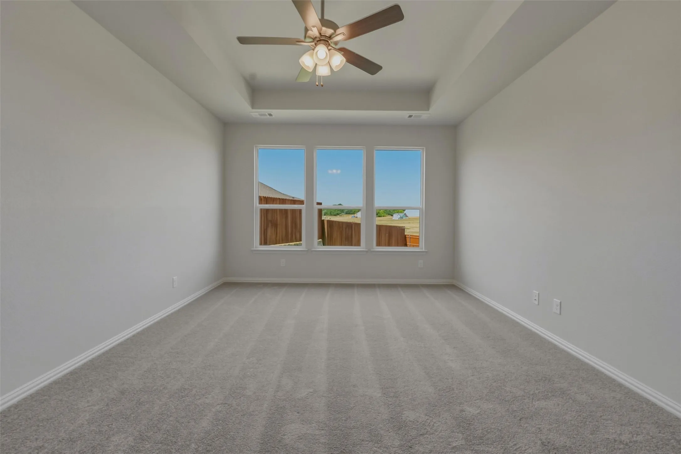 Spacious room featuring a tray ceiling, ceiling fan with light fixture, three large windows, light gray walls, and gray carpet flooring