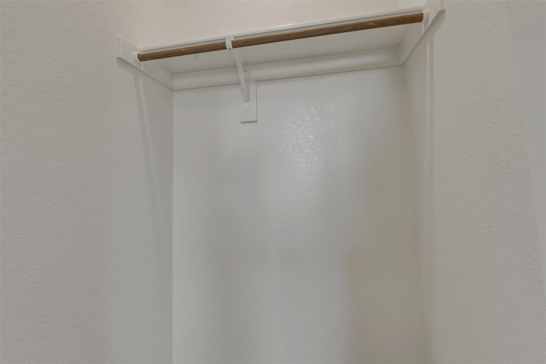 Recessed closet space featuring a natural wood-finish hanging rod and white metal supports, set against light-toned textured walls