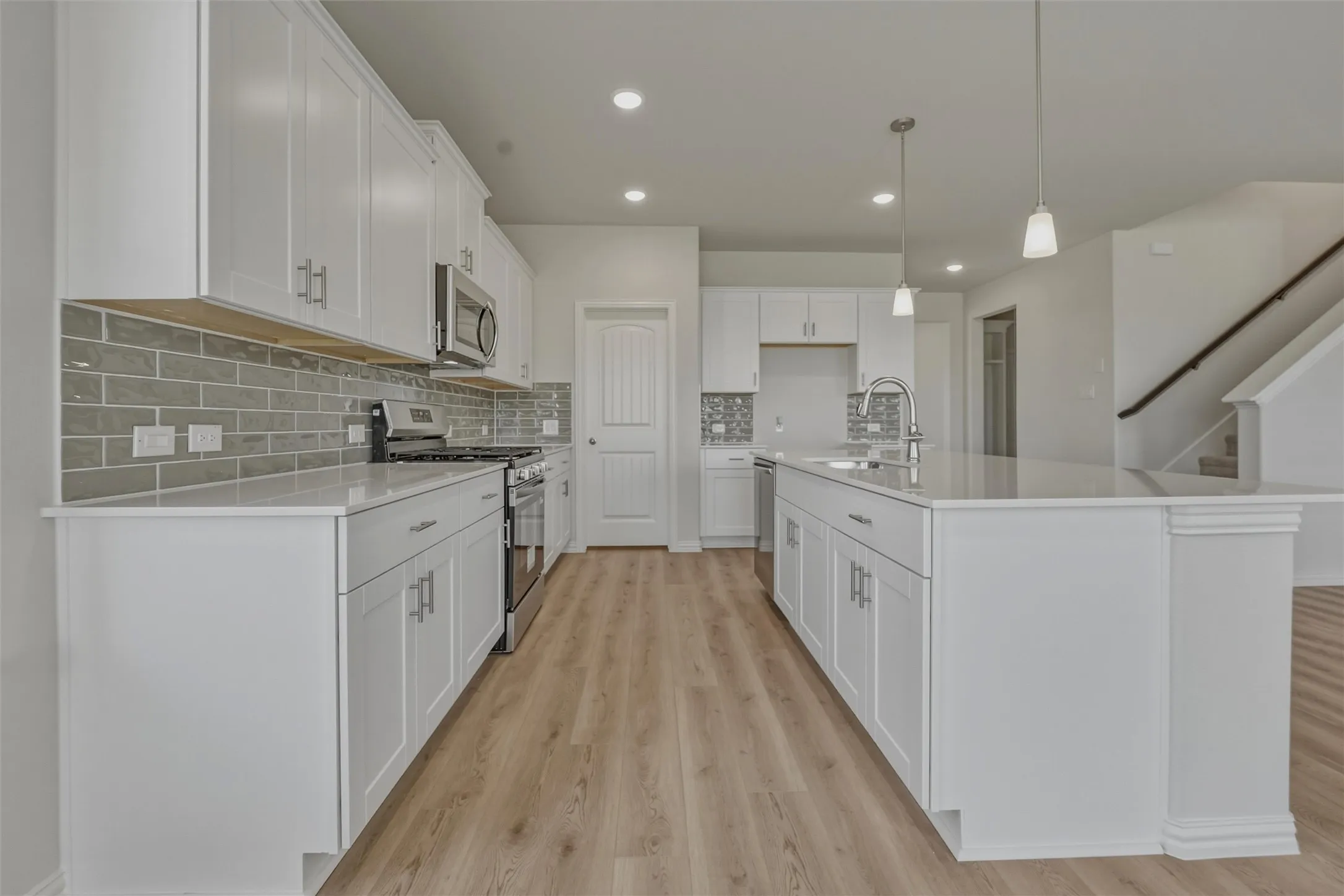 Modern kitchen featuring white shaker cabinetry, stainless steel appliances, a subway tile backsplash, light wood-finish flooring, and an expansive center island with pendant lighting