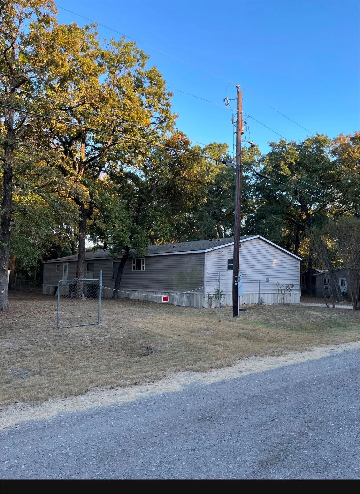 Single-story residence with light gray siding, perimeter fencing, mature trees, and a gravel road frontage