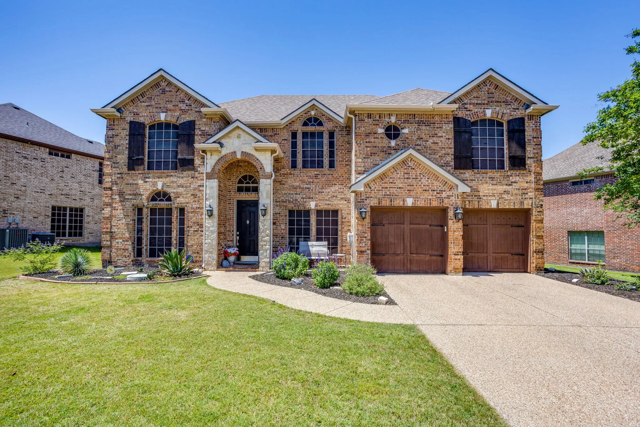 Brick facade residence featuring a covered entry, arched window details, dark shutters, a two-car garage with wood-finish doors, and a concrete driveway