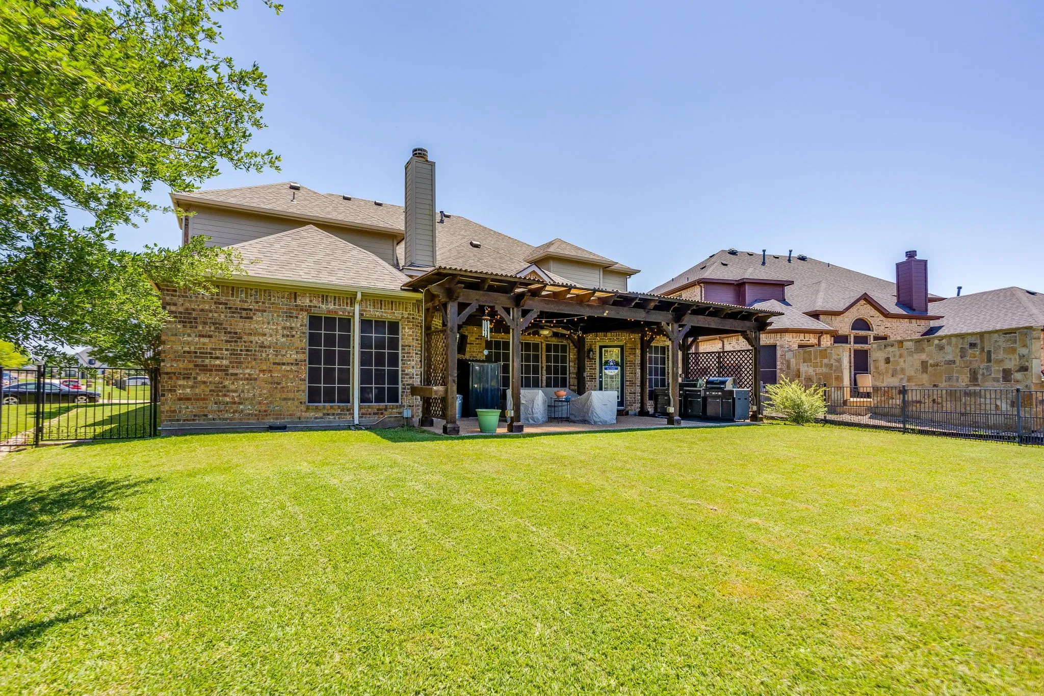 Spacious backyard featuring a large pergola-covered patio, extensive lawn, brick and stone exterior, and a wrought iron perimeter fence