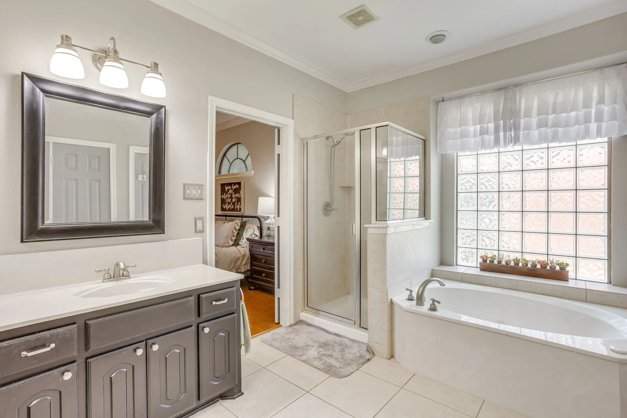 Bathroom featuring a large soaking tub, glass block window, walk-in shower with clear glass enclosure, single vanity with dark cabinetry, and tile flooring
