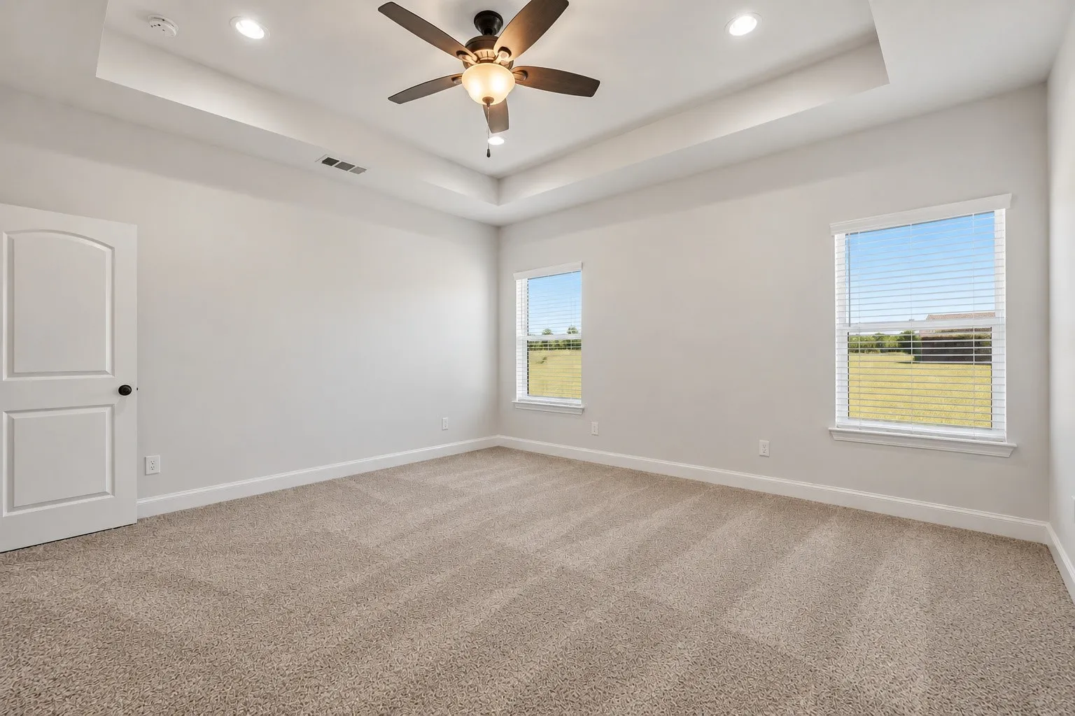 Bright and spacious bedroom featuring neutral carpeting, a recessed ceiling with ceiling fan and integrated lighting, and dual windows offering natural light and pastoral vistas