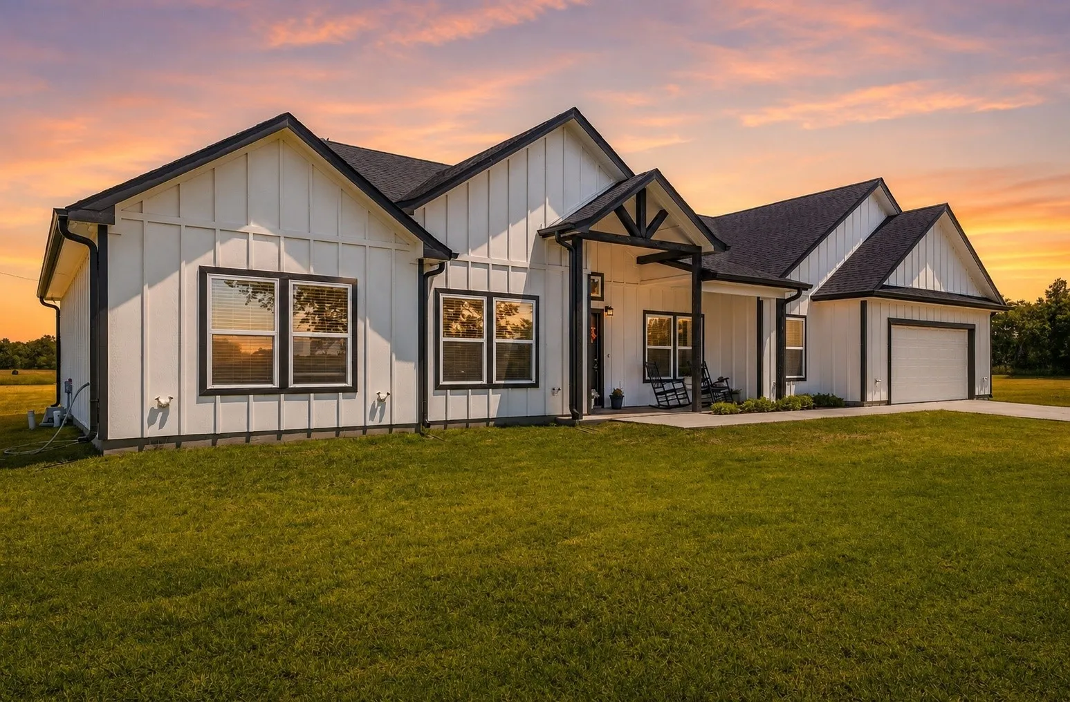 Modern farmhouse exterior featuring white board and batten siding, a dark asphalt shingle roof, black trim windows, a covered front porch, and an attached garage with a white panel door