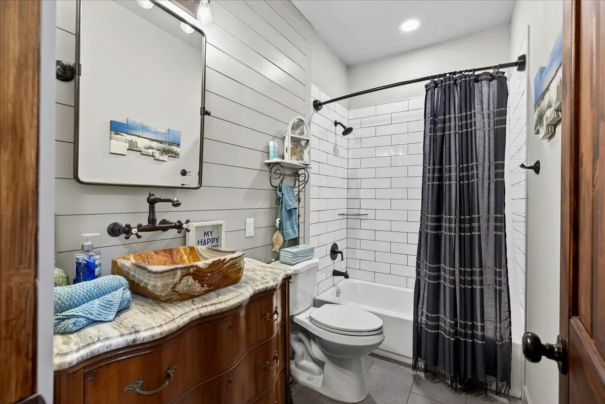 Hall Bathroom on Level 1 featuring a unique natural stone vessel sink, wall-mounted faucet, and a vanity with a light-colored countertop, shiplap accent wall, subway tile in shower/tub, gray tile floor.